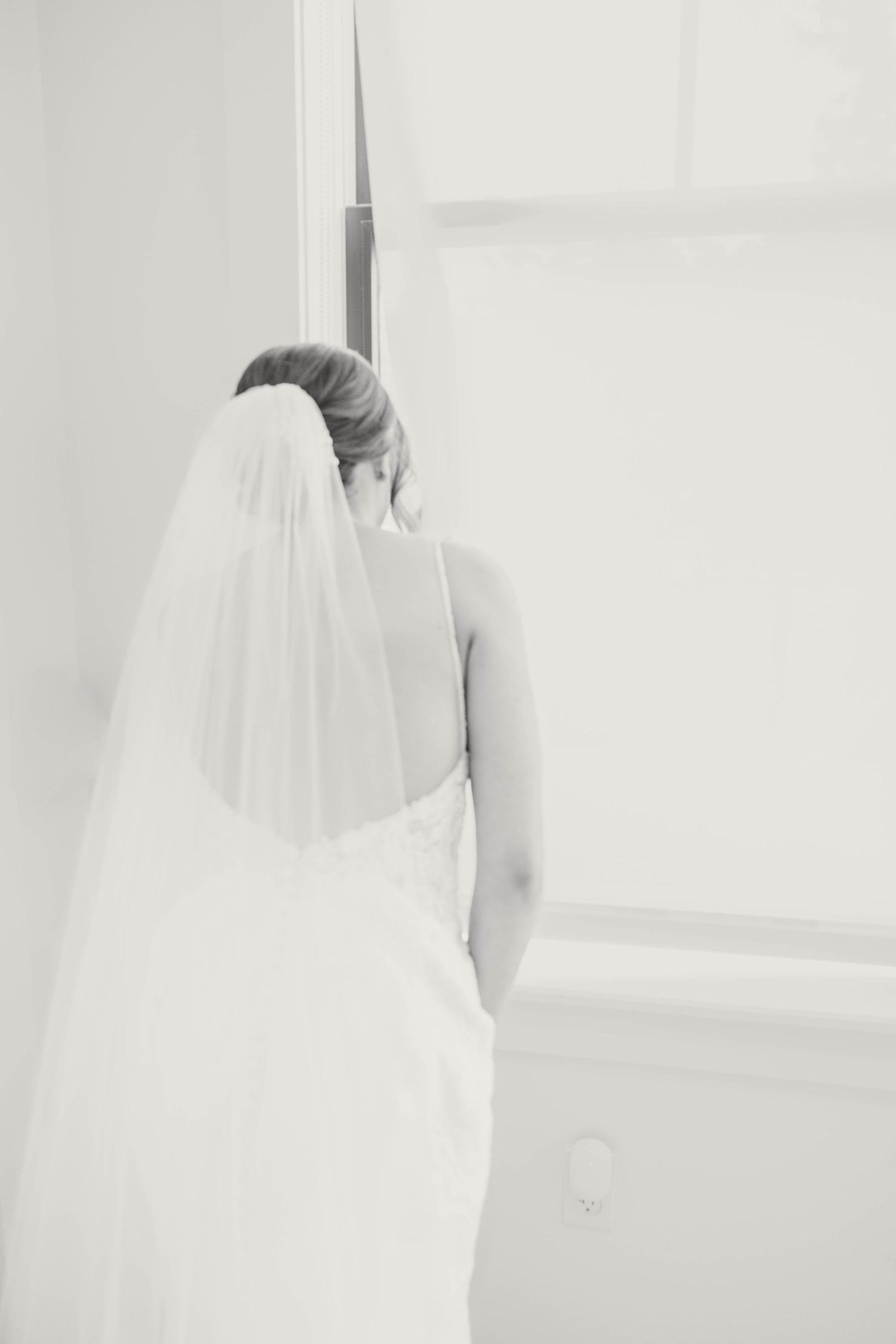 Black and white photo of bride in lace wedding gown with veil standing by white door.