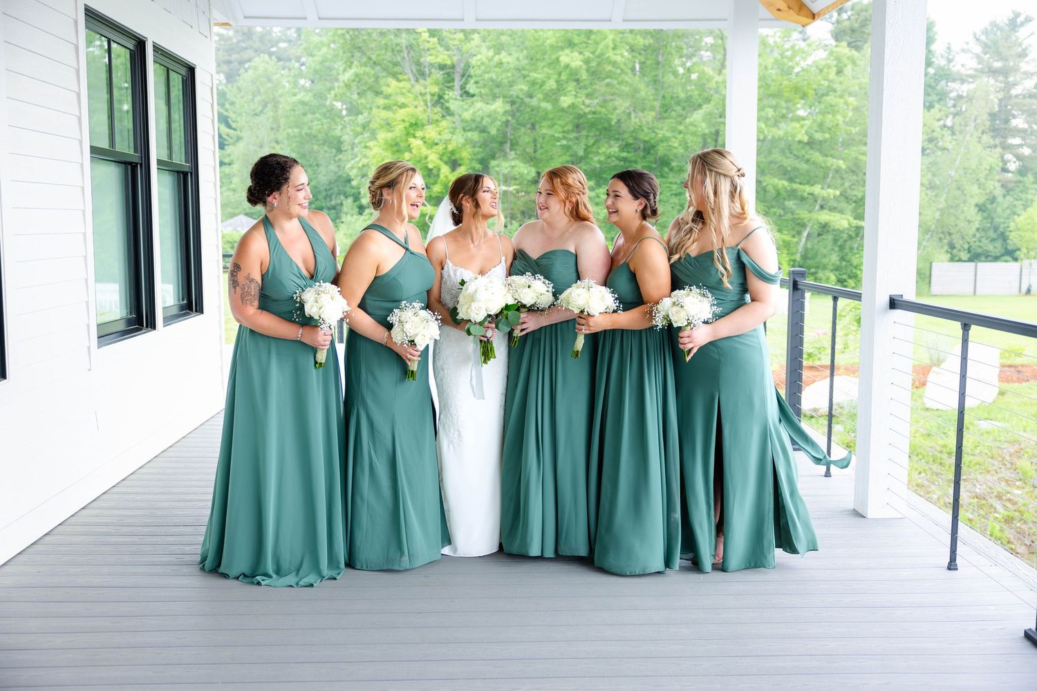 Bride and bridesmaids in elegant sage green dresses laugh together on wedding venue porch.
