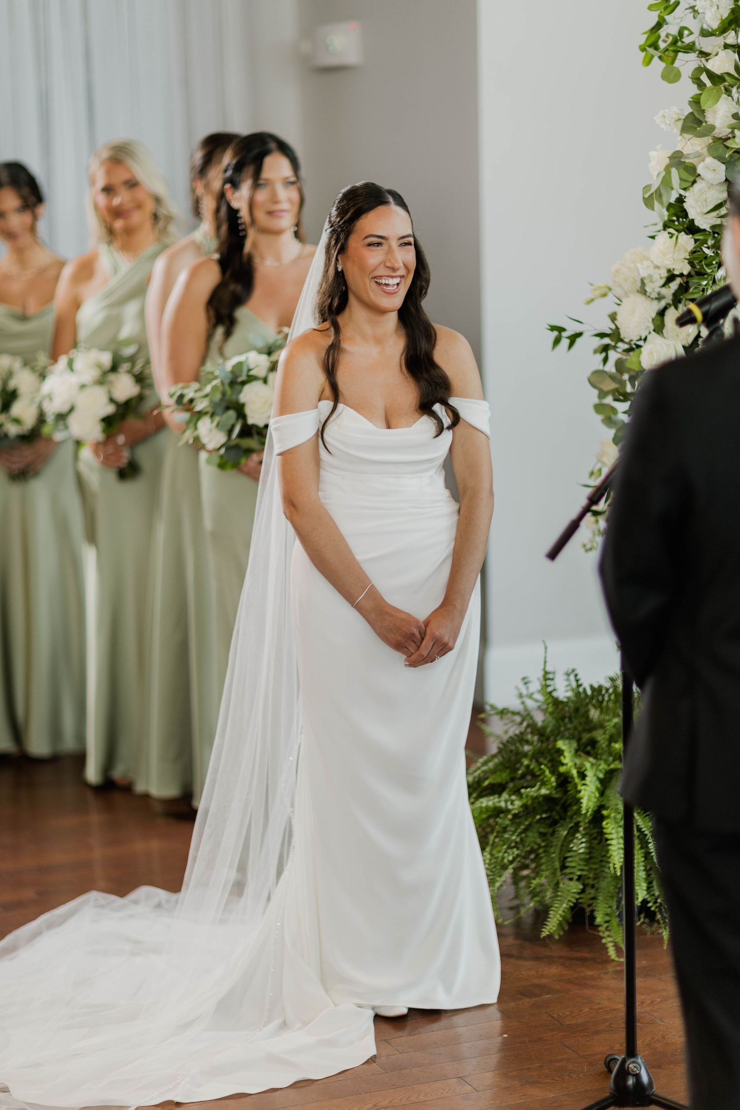 Smiling bride in white off-shoulder gown stands at altar with bridesmaids in sage green dresses holding white bouquets.