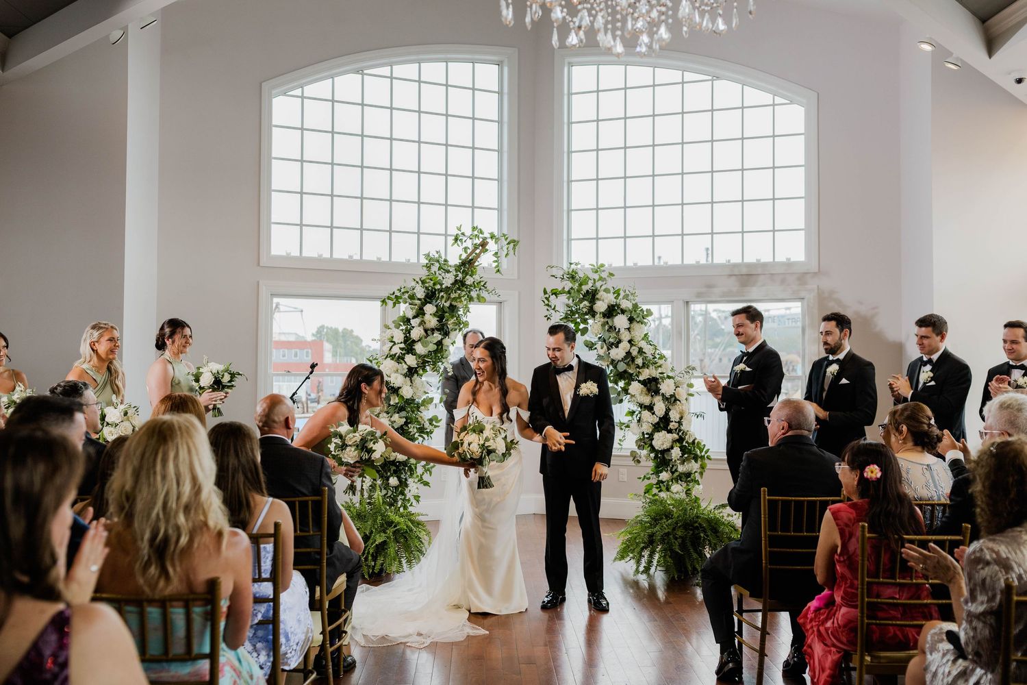 Bride and groom exchange vows under floral arch in bright white wedding venue.