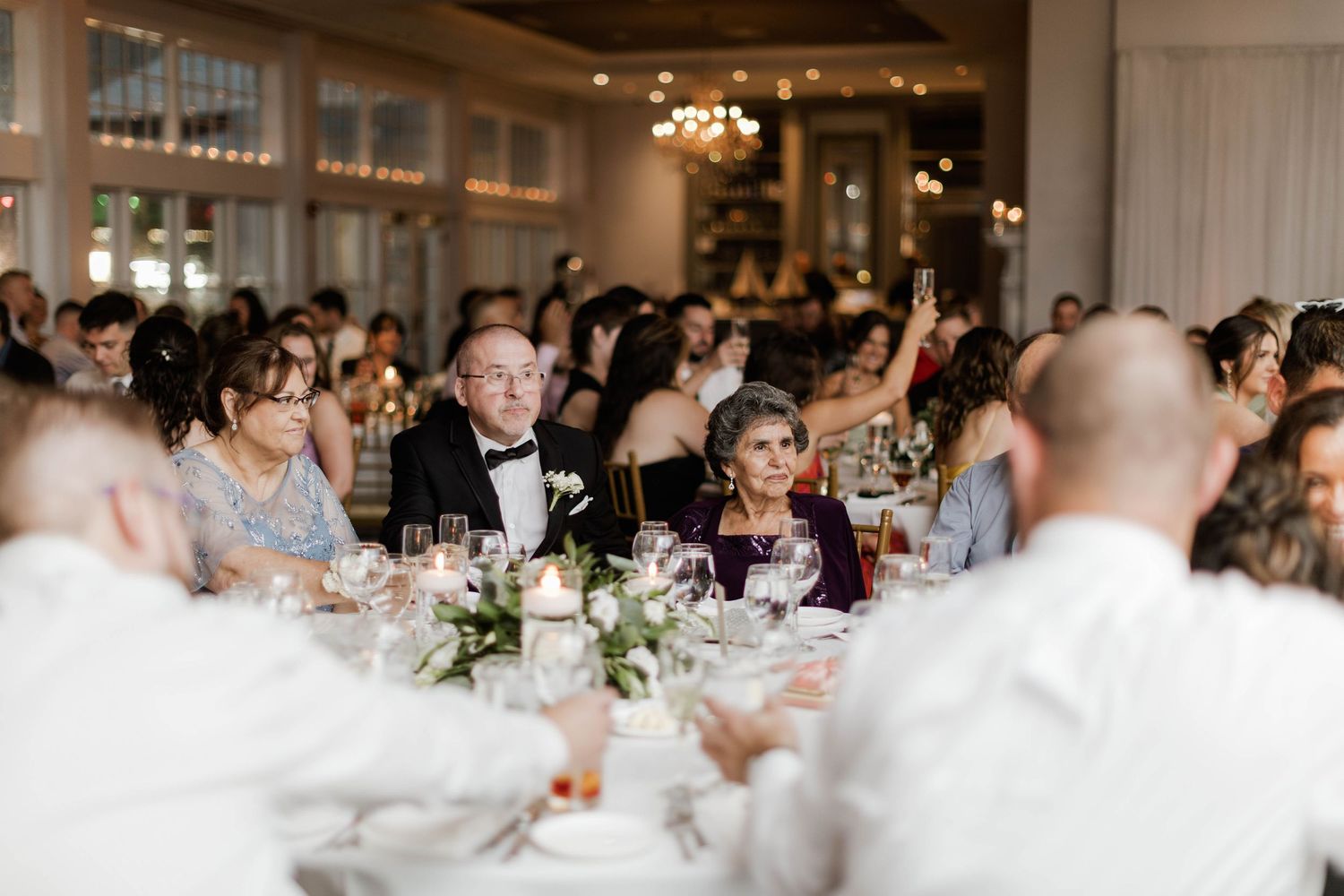 Elegantly dressed wedding guests seated at candlelit reception tables in a grand ballroom raise glasses.