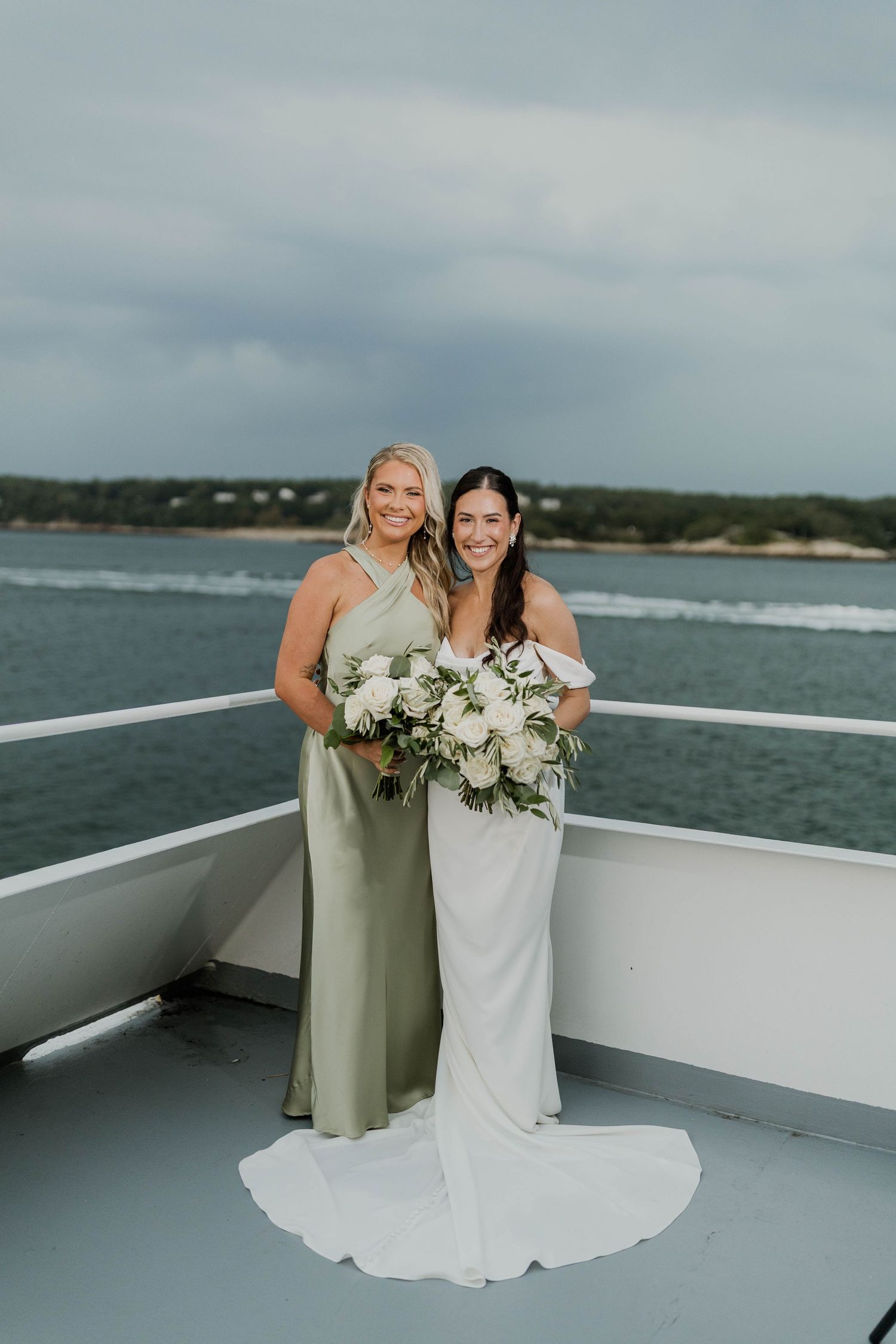Bride in white gown and bridesmaid in sage green satin dress posing on a boat with ocean views.