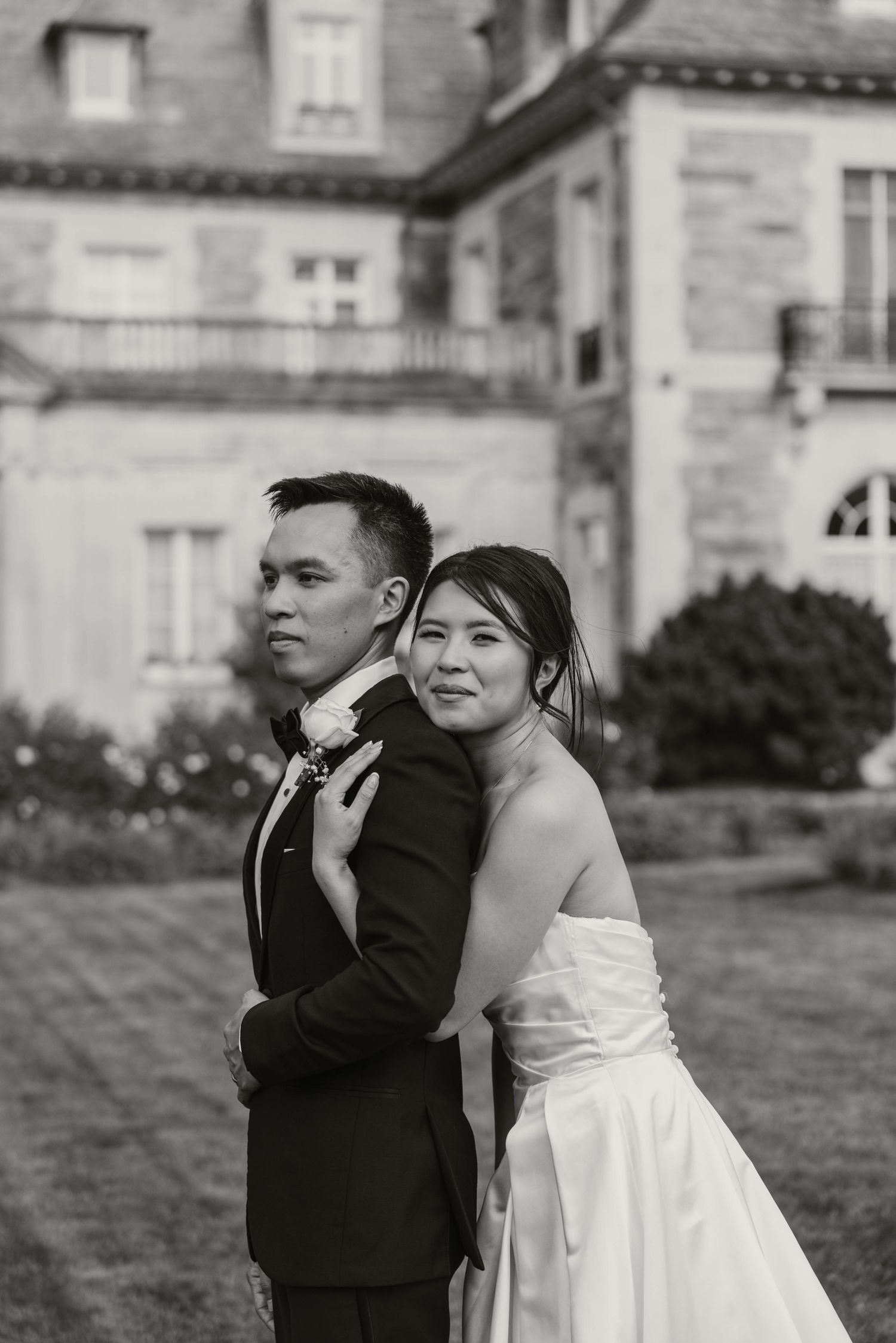 Black and white portrait of bride and groom embracing outside a stone mansion wedding venue.