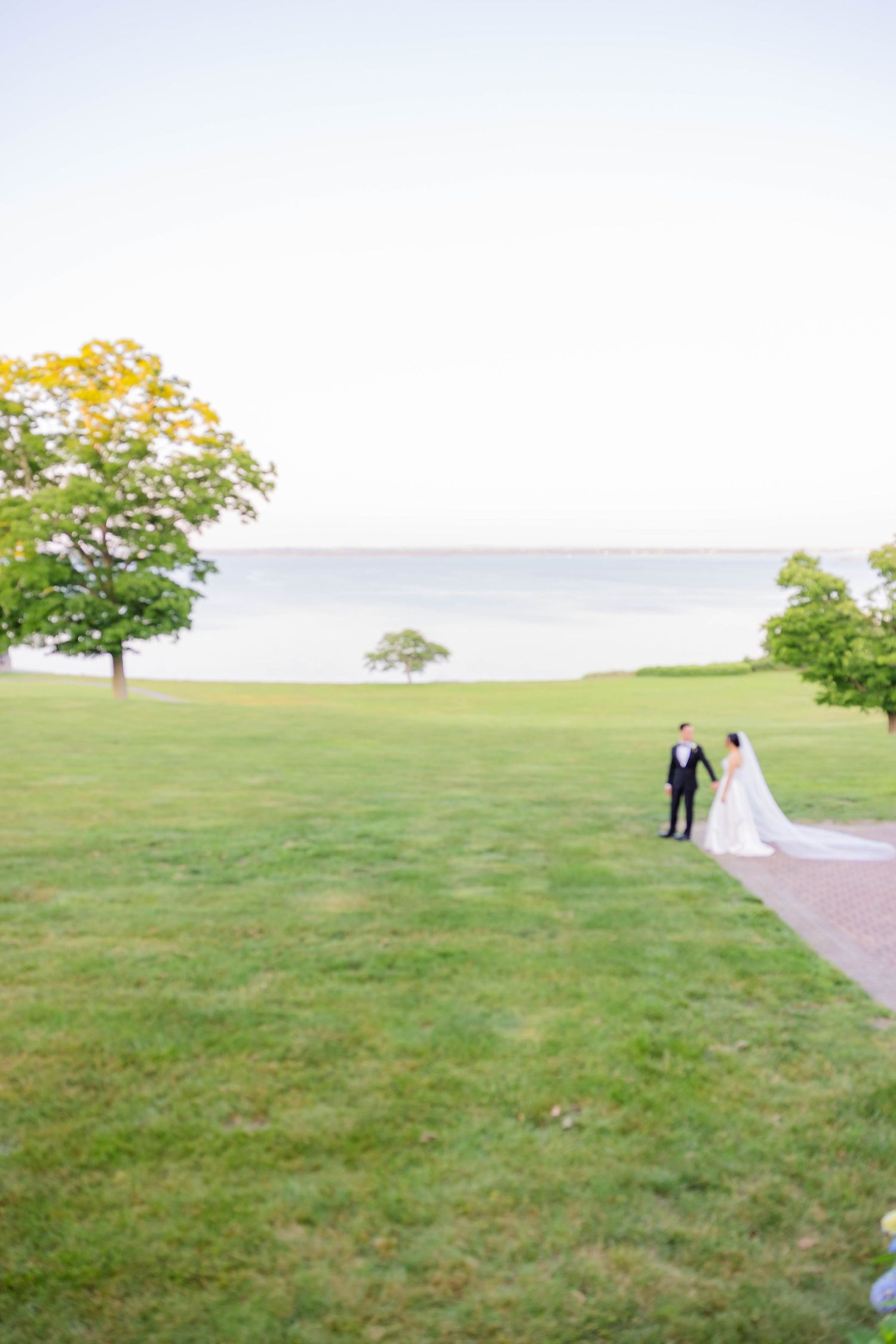 Bride and groom walk hand in hand across lush green lawn beside serene waterfront.