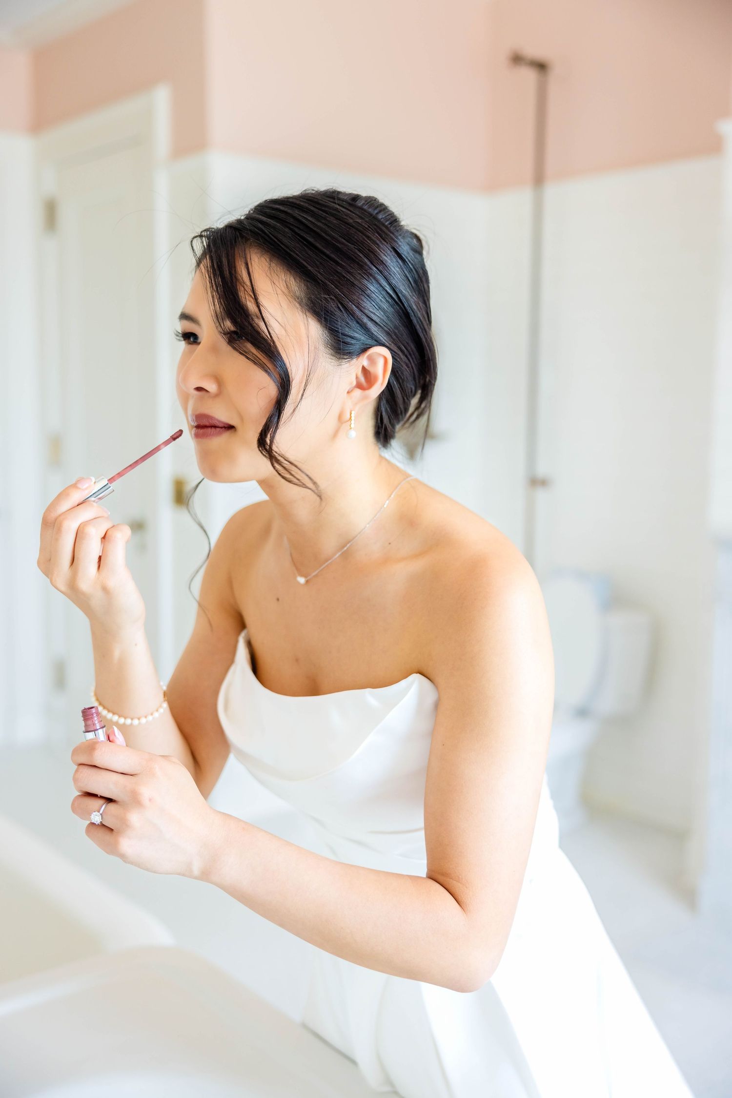 Asian bride applies nude lip gloss in bathroom mirror while wearing strapless white wedding gown and pearl bracelet.