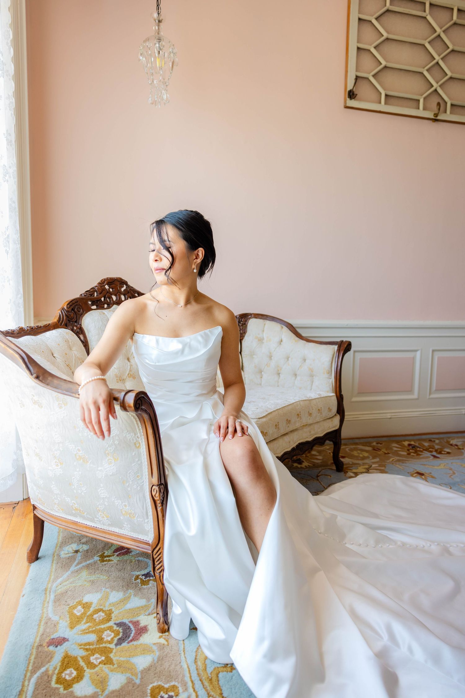 Bride in strapless white gown sitting on vintage sofa laughing joyfully in elegant pink room.