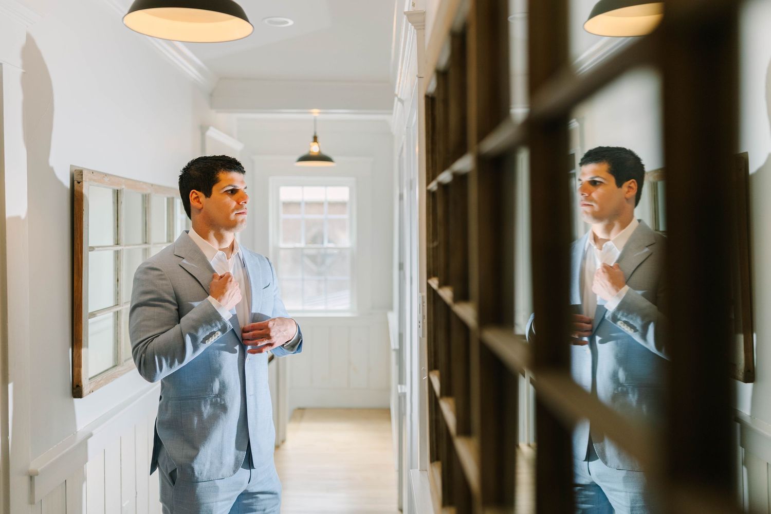 A well-dressed man in a gray suit adjusts his tie while standing in a bright, elegant hallway with a mirror reflection.