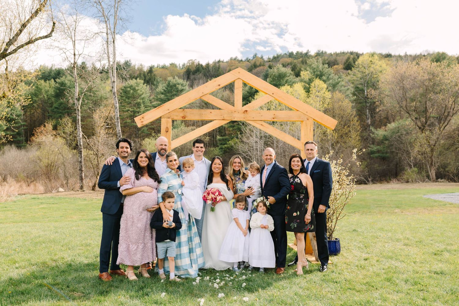 Wedding family group portrait under wooden arch with colorful autumn hillside backdrop on green lawn.