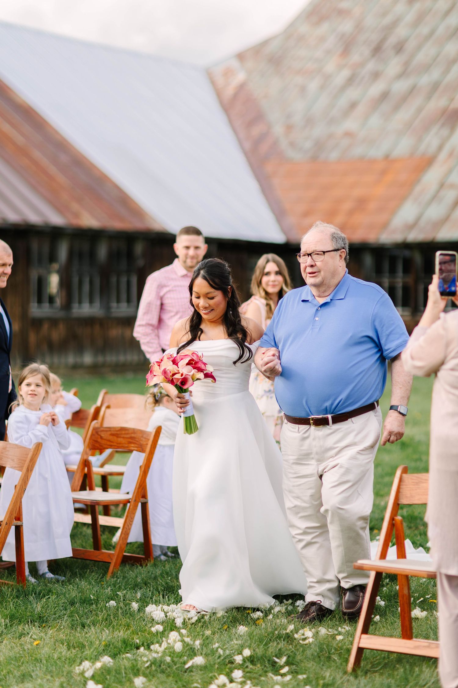 Bride in white gown walks down outdoor aisle with older man past wooden chairs at rustic barn wedding.
