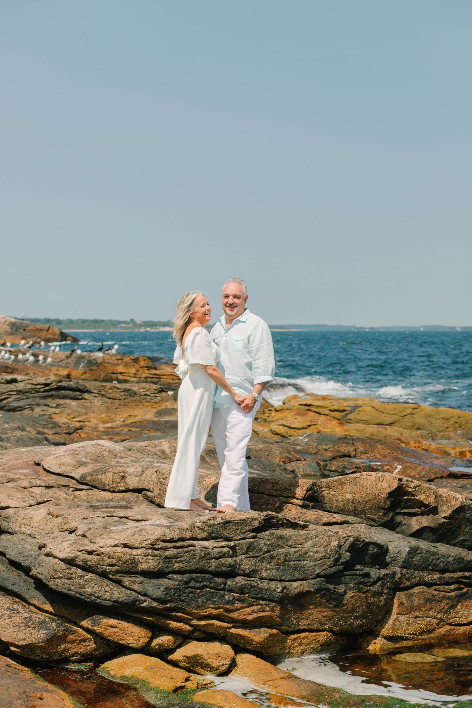 Senior couple holding hands and smiling on rugged coastal rocks with blue sea in the background.