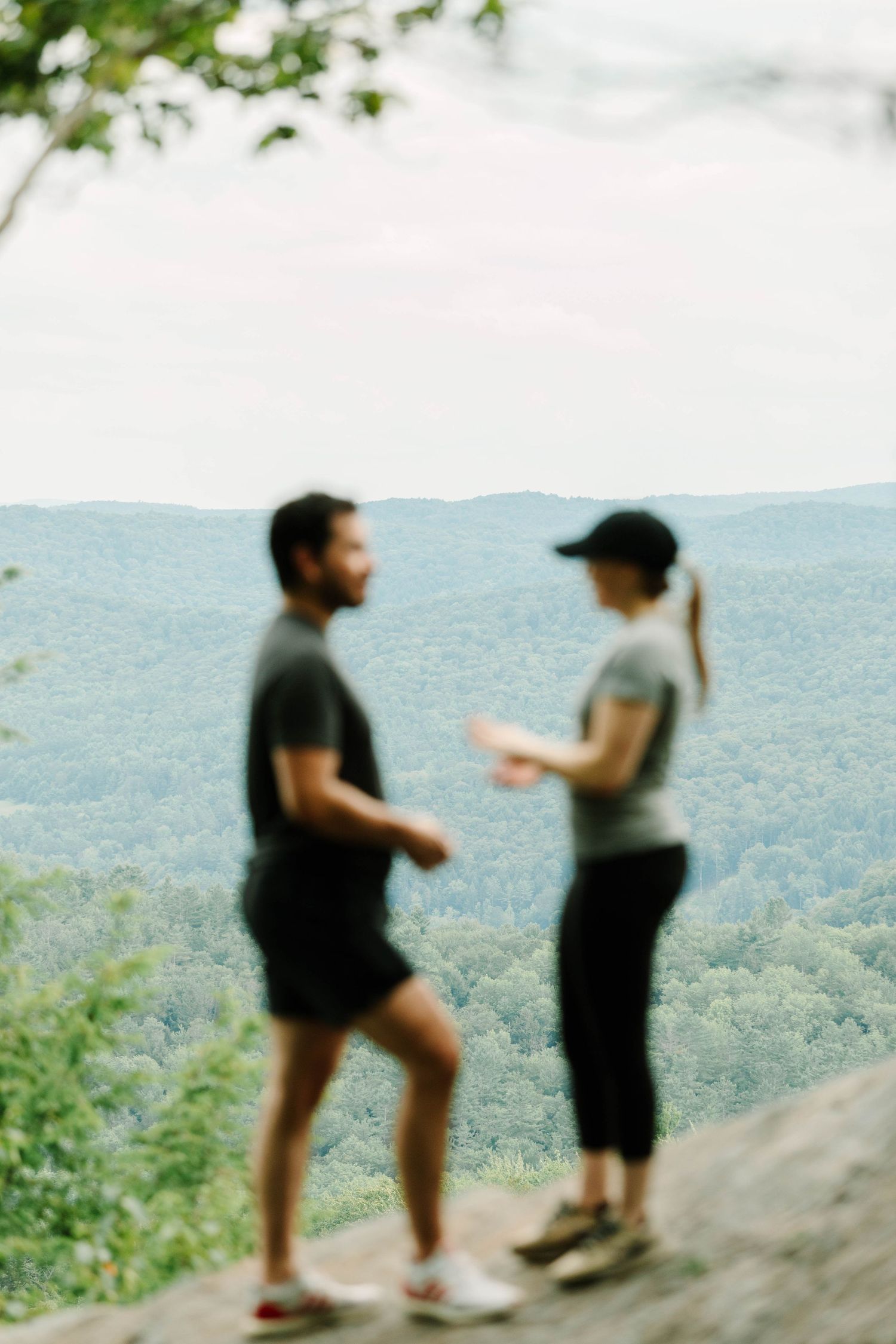 Blurred couple standing on a rocky overlook with expansive forested mountain landscape behind them.