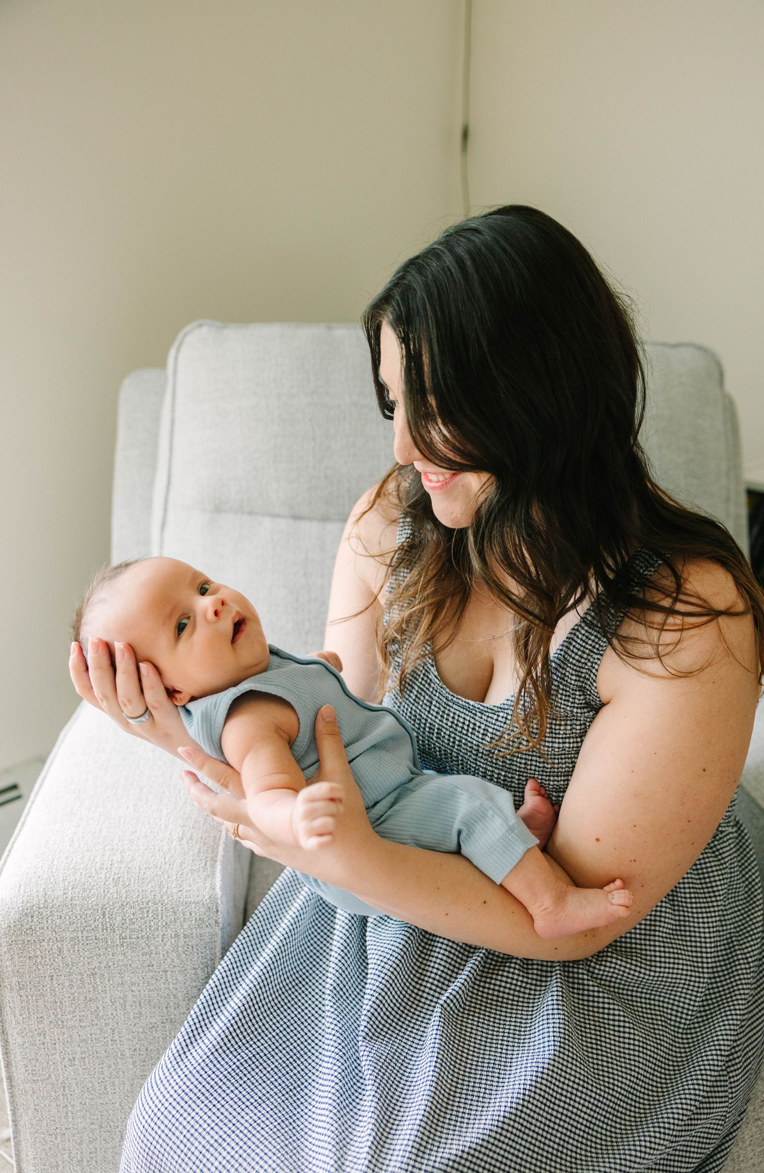 A smiling dark-haired mother holds her newborn baby in a blue onesie while sitting in a gray chair.