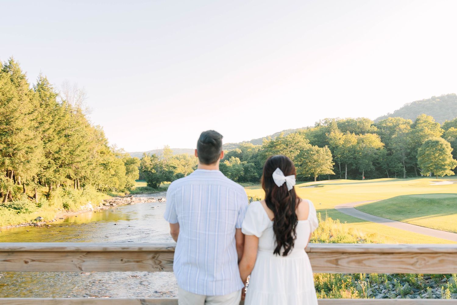 Couple holding hands on a bridge overlooking a scenic river and lush green hills on a sunny day.