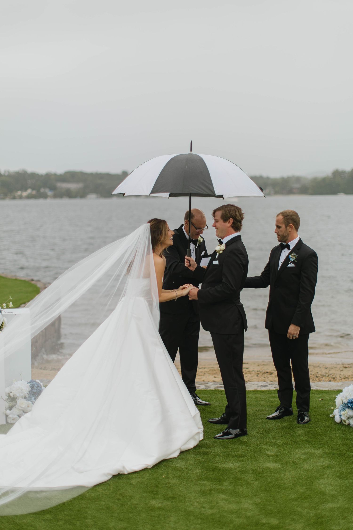 Outdoor waterfront wedding ceremony with bride in white gown and veil, groom, officiant, and groomsman under black umbrella.