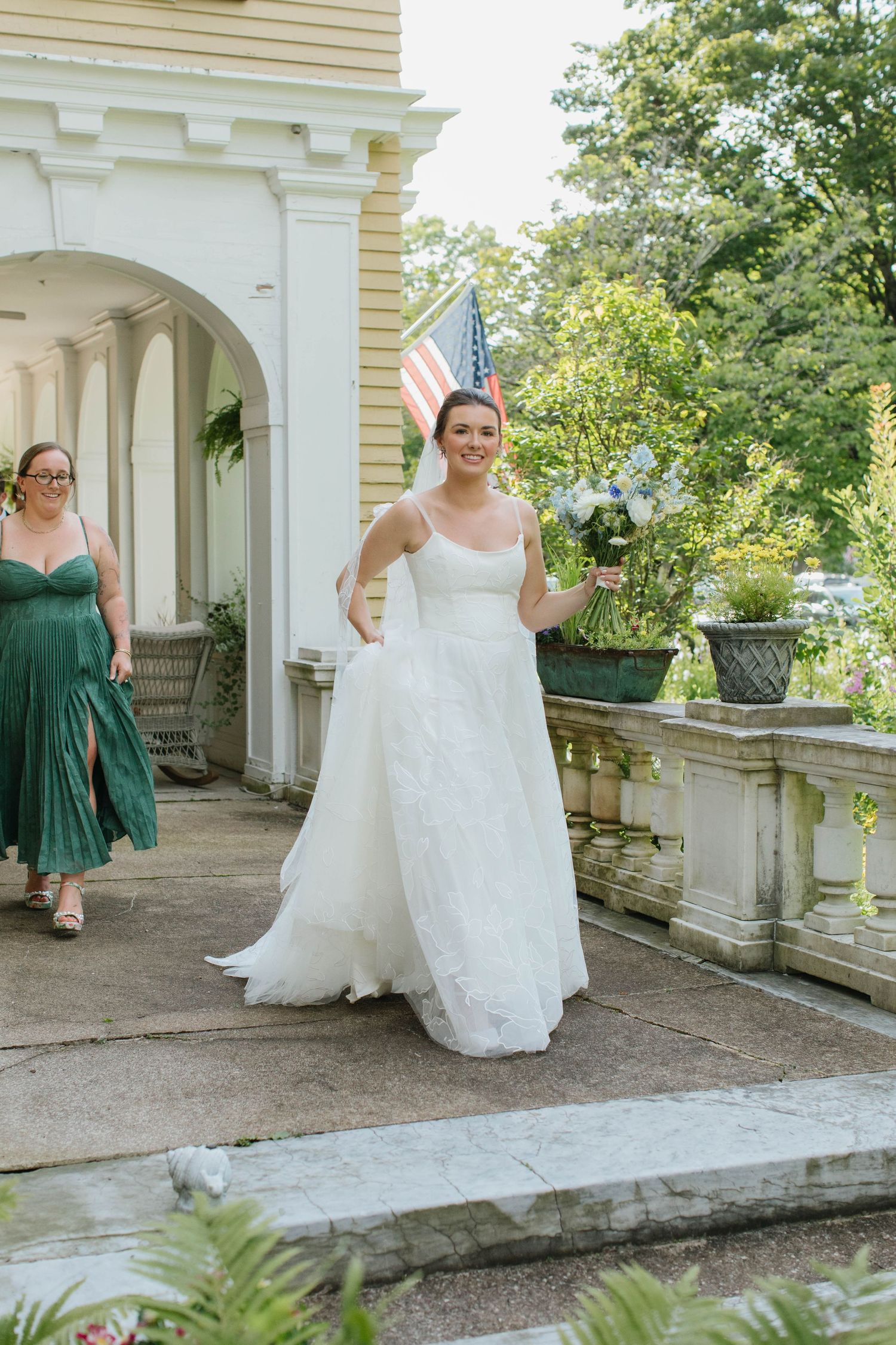 Elegant bride in white A-line gown walks down steps at classic venue with bridesmaid.