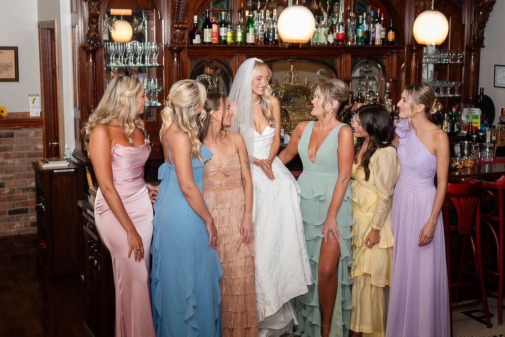 Bride surrounded by bridesmaids in colorful gowns laughing together at a bar during wedding celebrations.