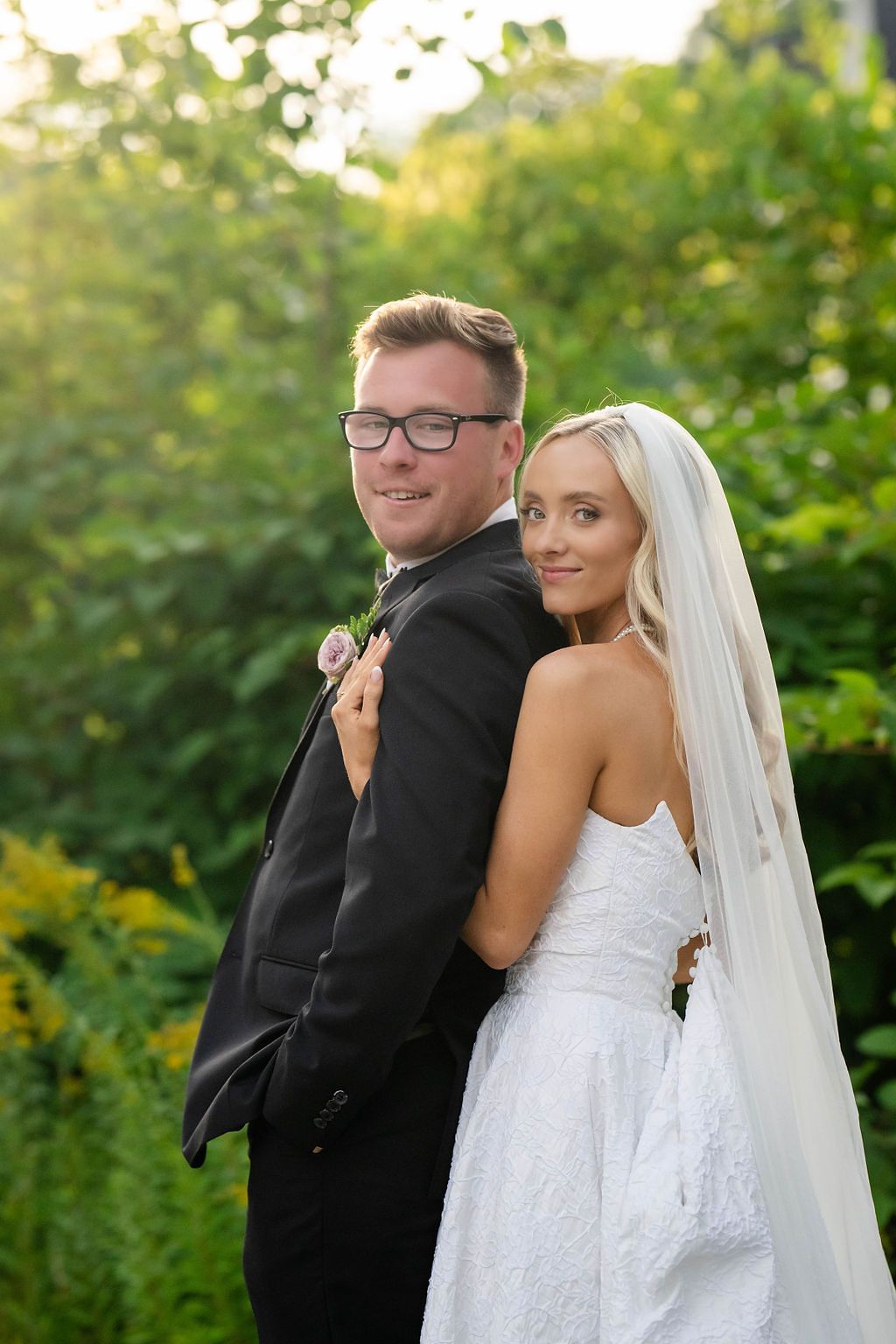 Bride and groom posing together in lush green garden, bride in white lace gown with veil, groom in black tux.