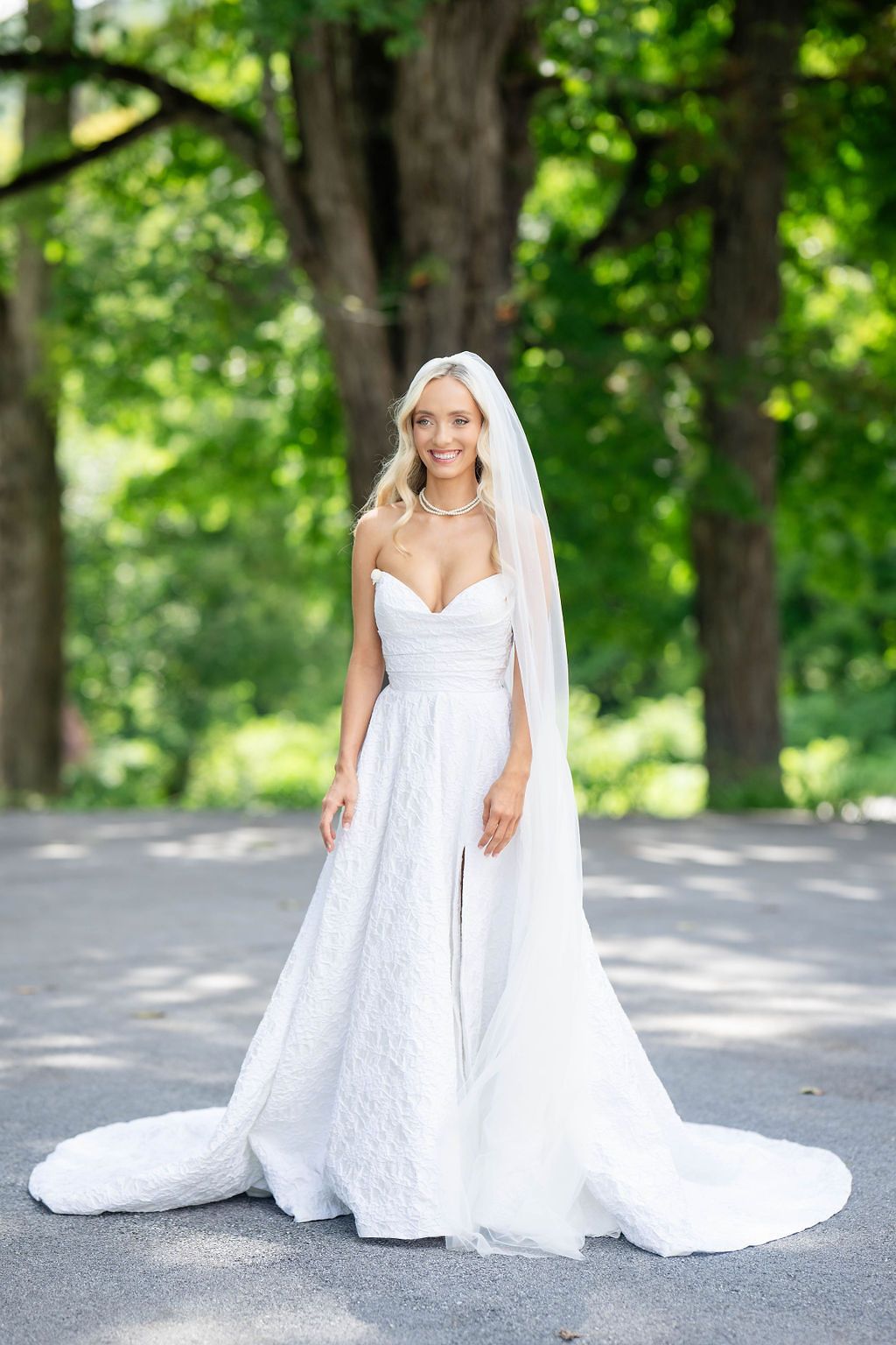 Radiant bride in strapless white lace ball gown and cathedral veil stands outdoors surrounded by lush green trees.