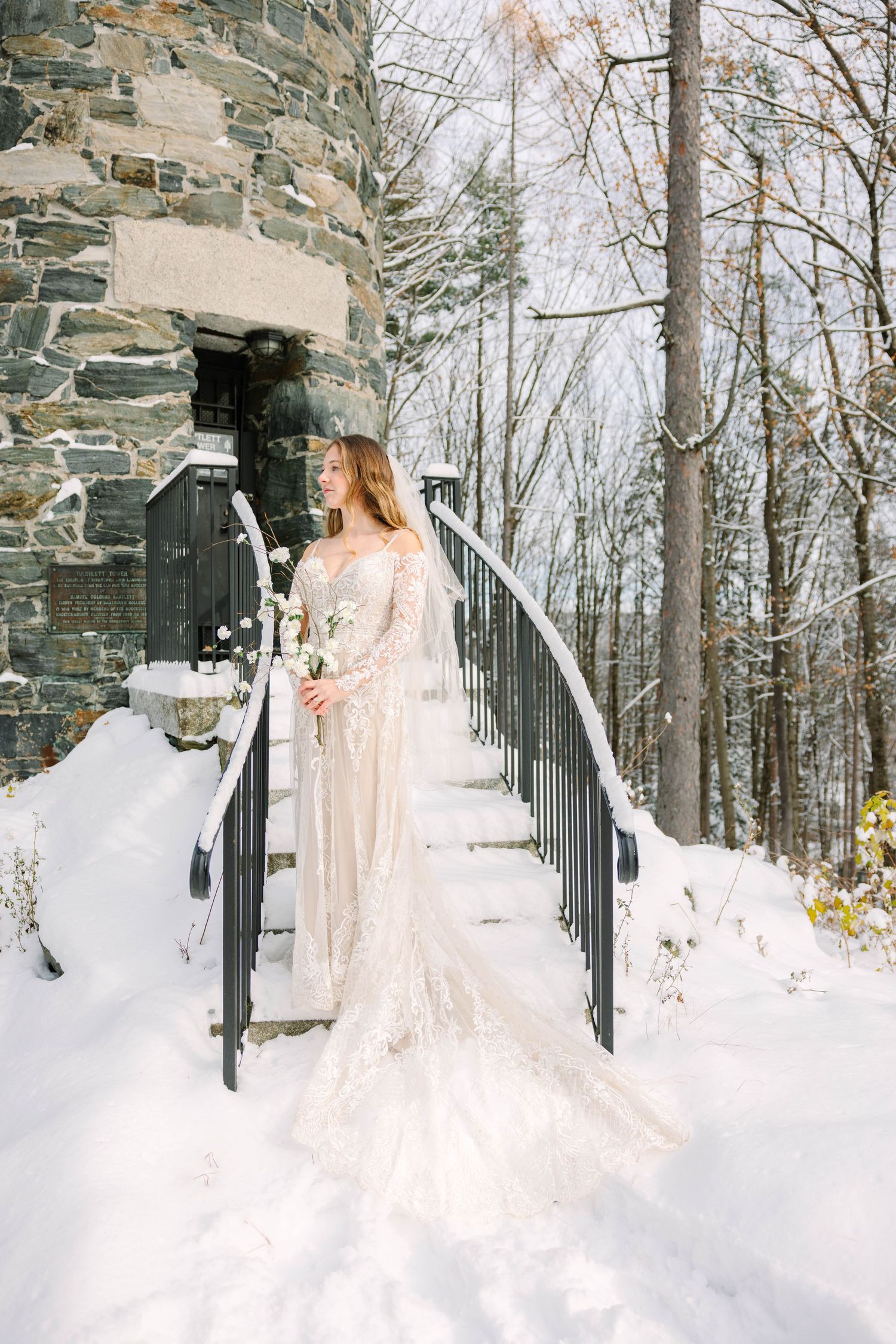 Elegant bride in lace wedding dress stands on snowy stone building staircase holding white flowers.