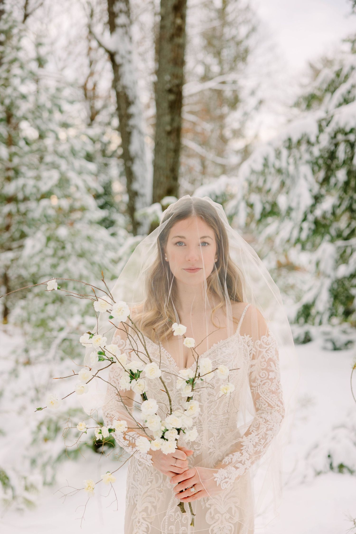 Bride in lace gown with sheer veil holding white floral branch in snow-covered winter woods.
