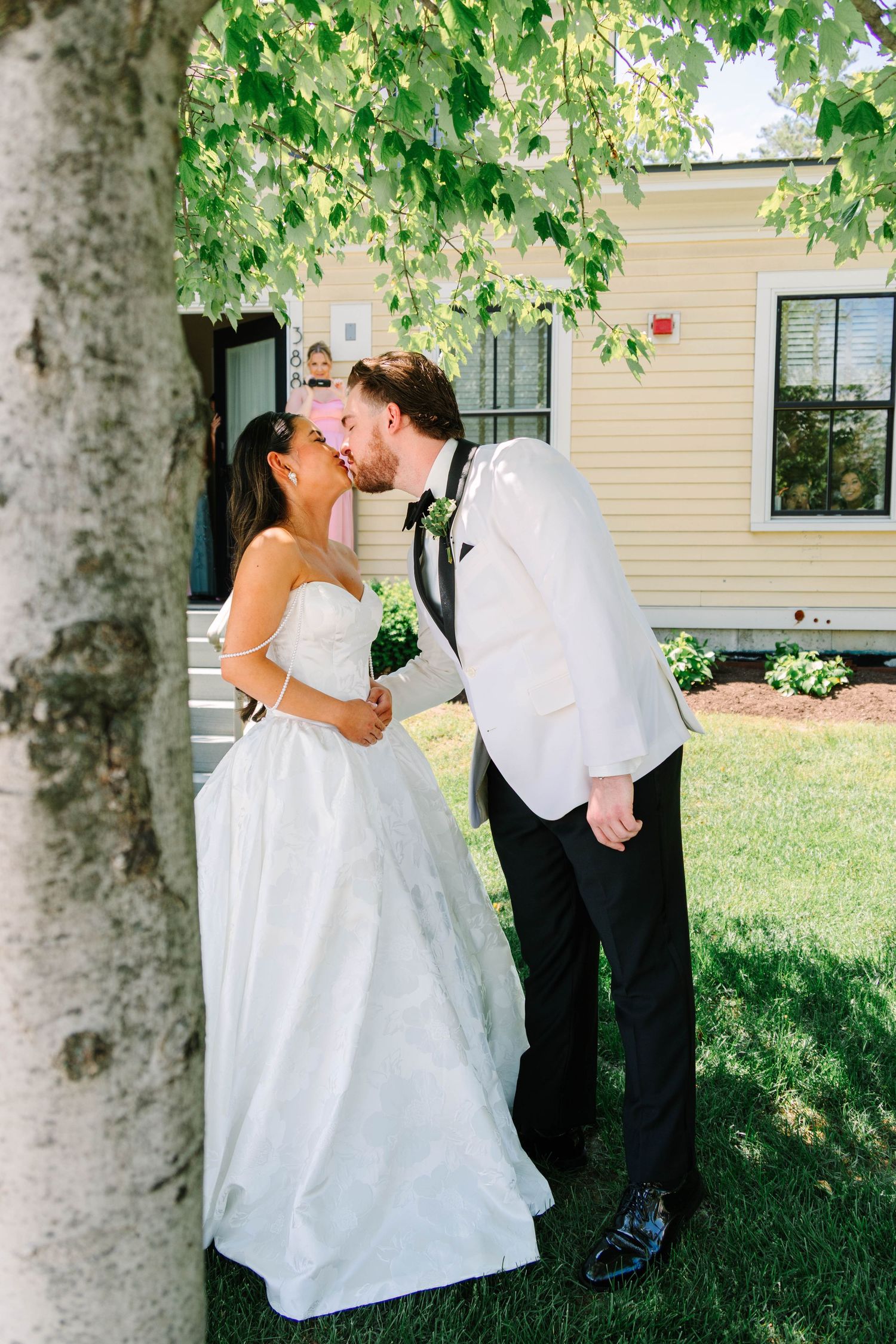 Bride and groom sharing first kiss under lush green tree at outdoor summer wedding venue.