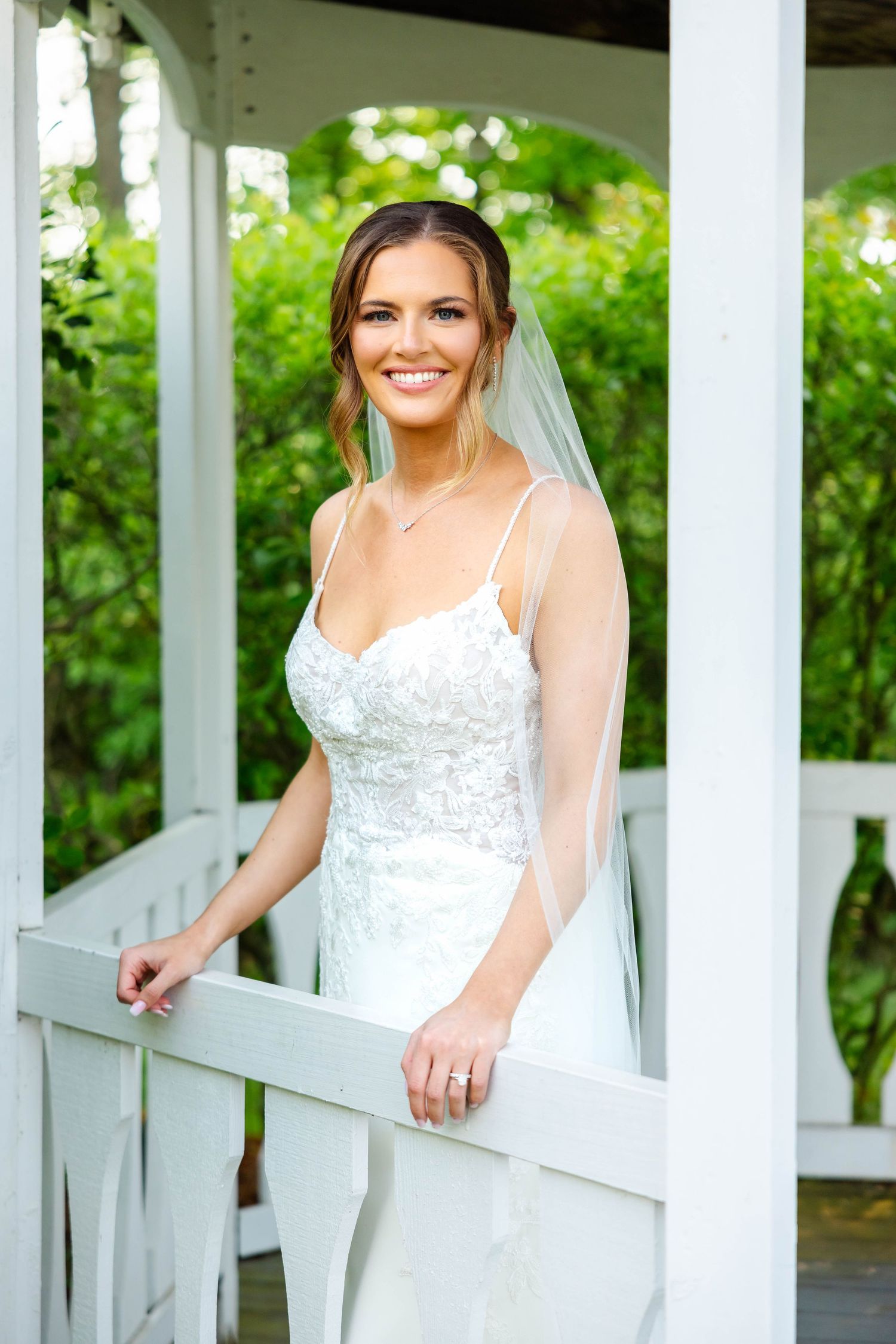 Radiant bride in lace spaghetti strap gown and veil posing in white garden gazebo.