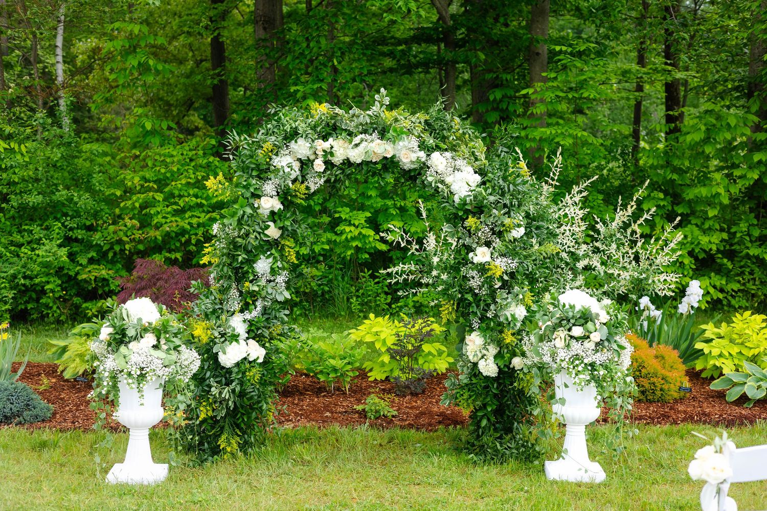 Elegant outdoor wedding arch adorned with lush white roses and greenery flanked by floral urns.