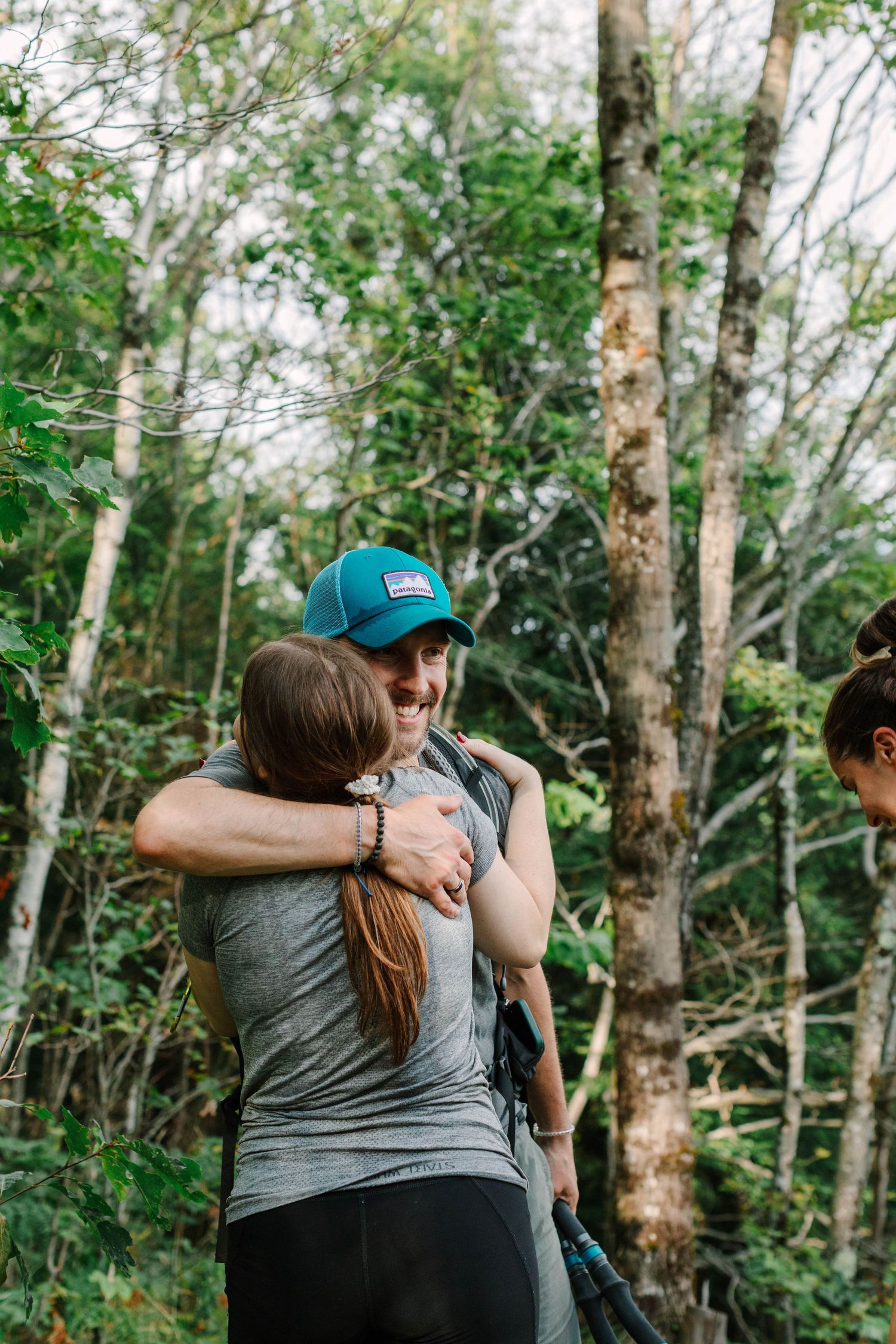 Man in blue cap joyfully hugging a woman in a gray shirt in a wooded forest setting.