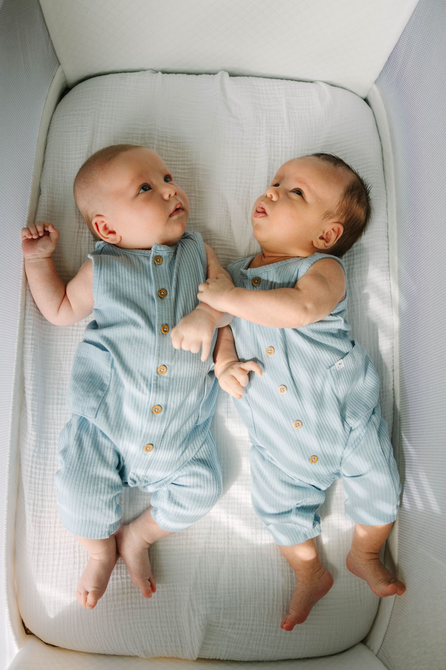 Twin newborn babies in matching light blue striped rompers lying together in a white bassinet.
