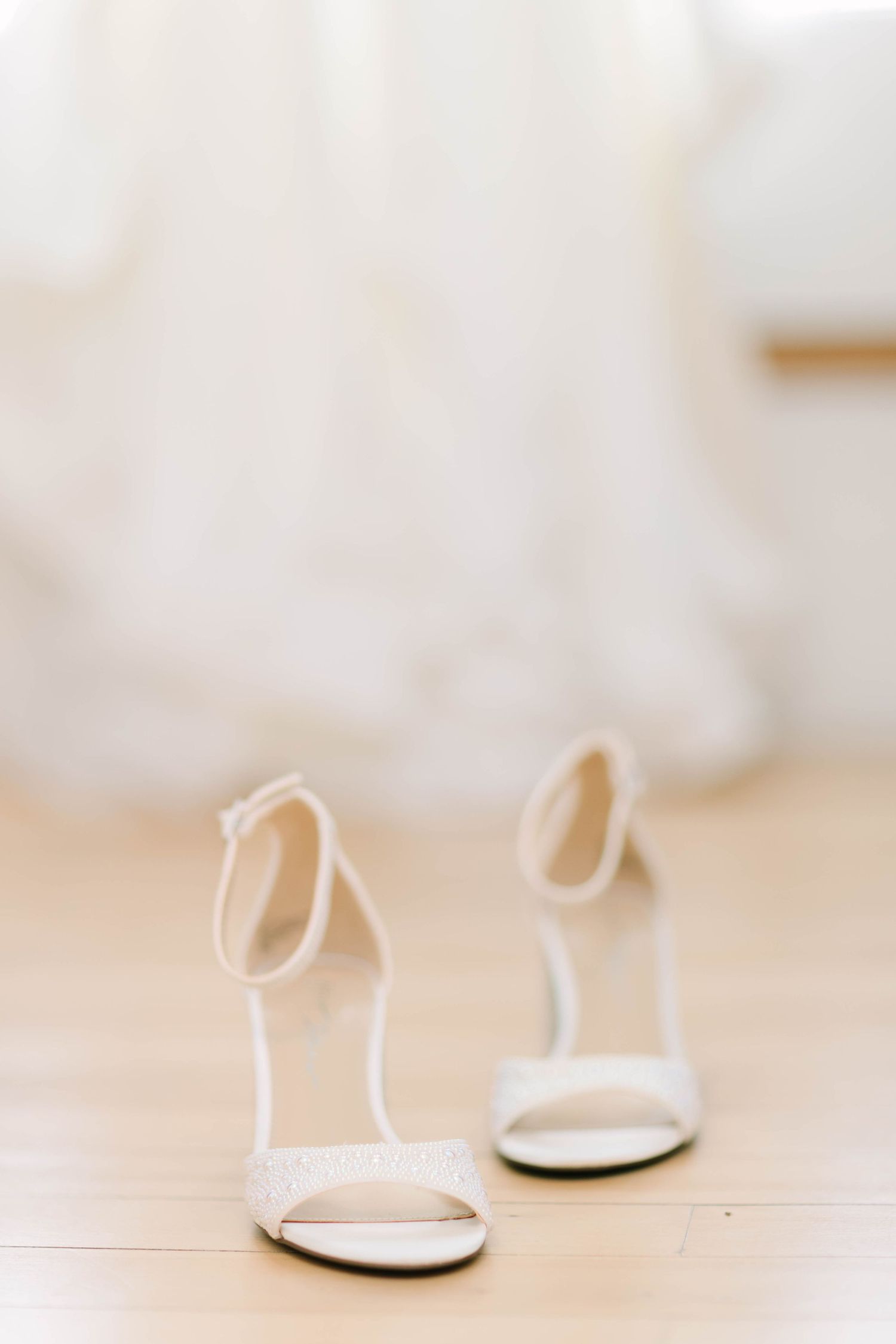 Elegant white bridal heels with ankle straps on a wooden floor, with a blurred wedding dress in the background.