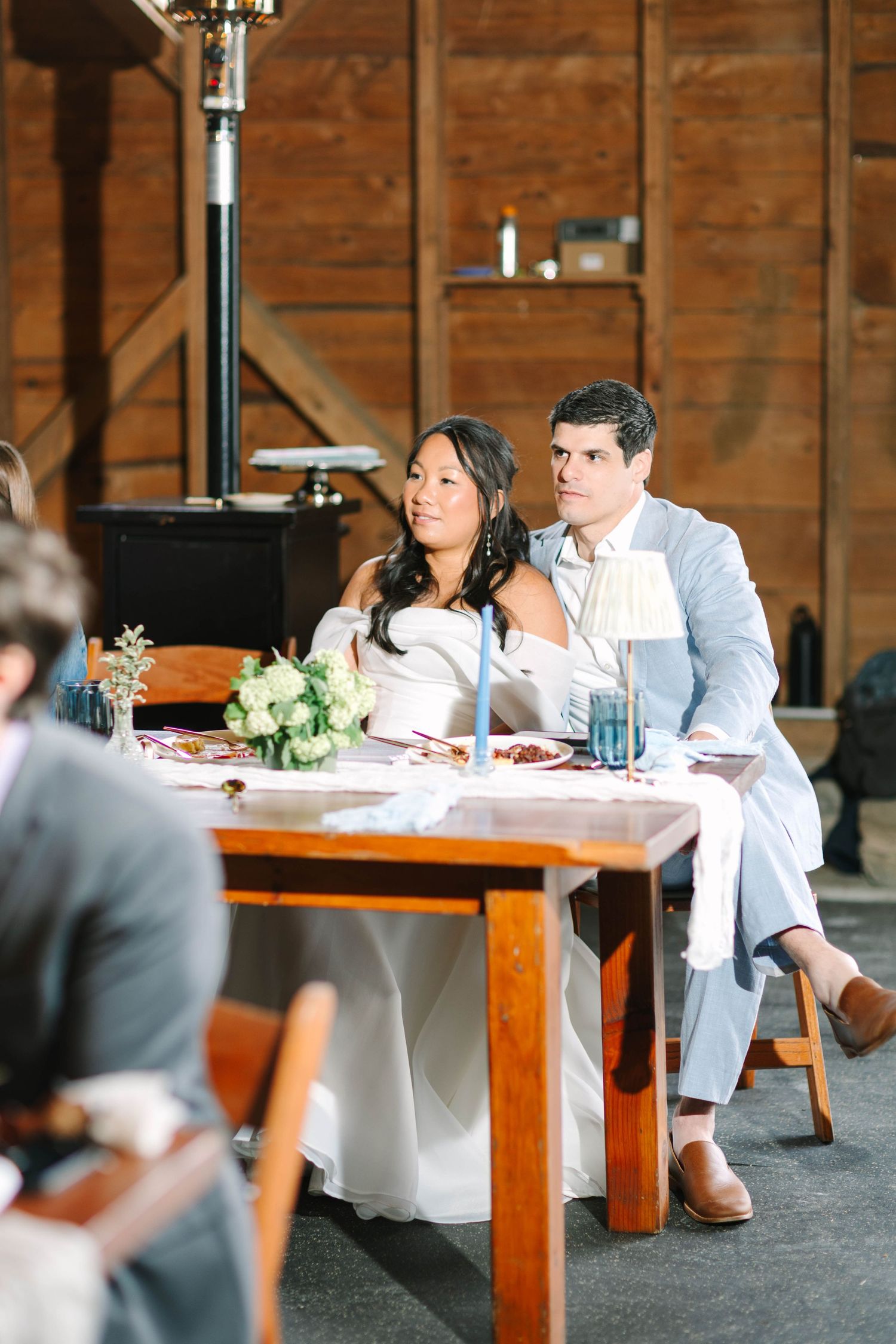 Bride and groom sit together at sweetheart table inside rustic barn during wedding reception speeches.