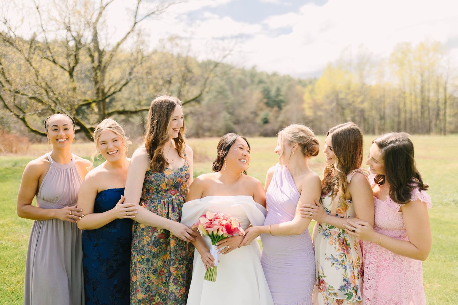 Smiling bride holding colorful bouquet surrounded by bridesmaids in mix of floral and lavender gowns outdoors.