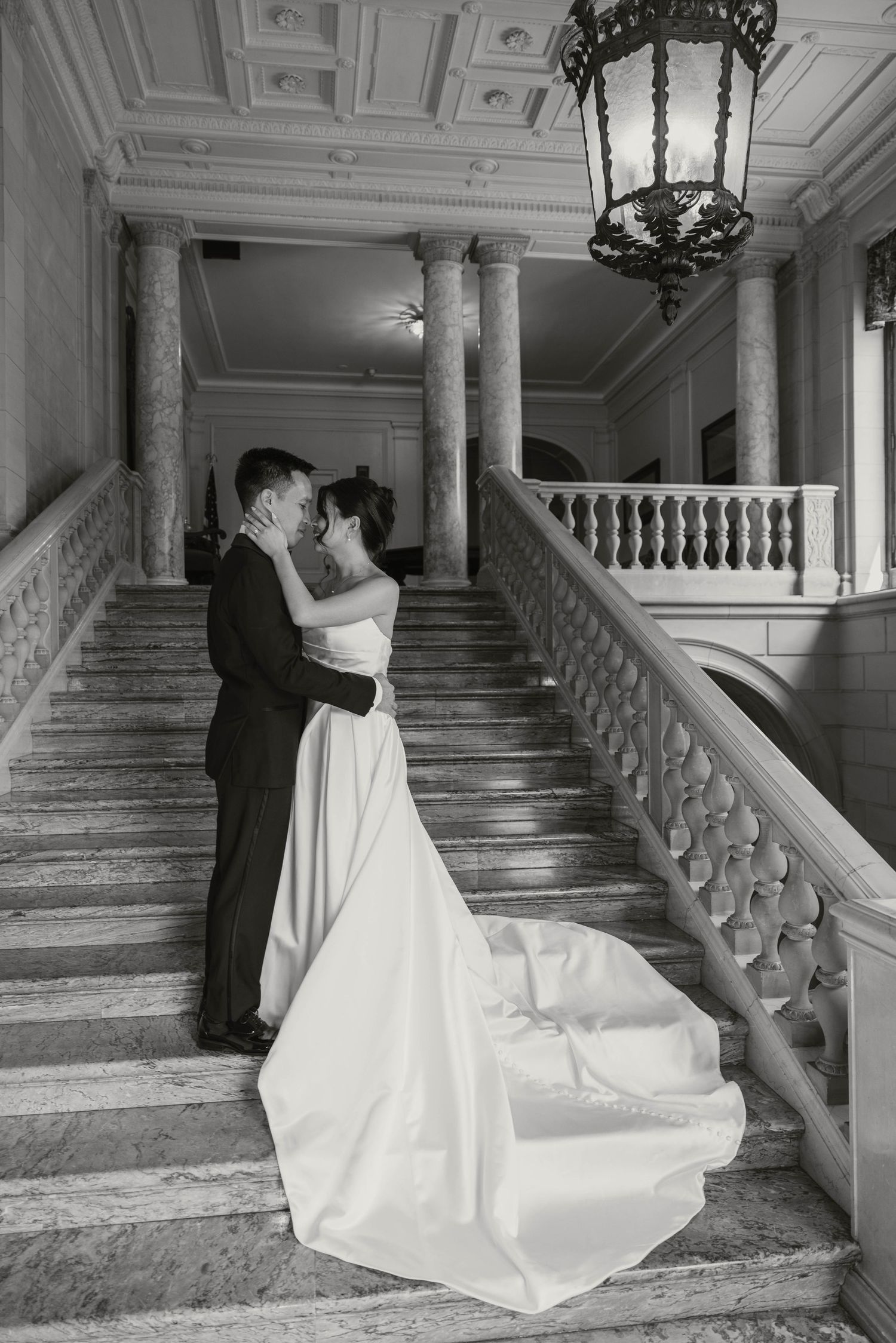Black and white photo of bride and groom embracing on grand marble staircase with ornate balustrade.