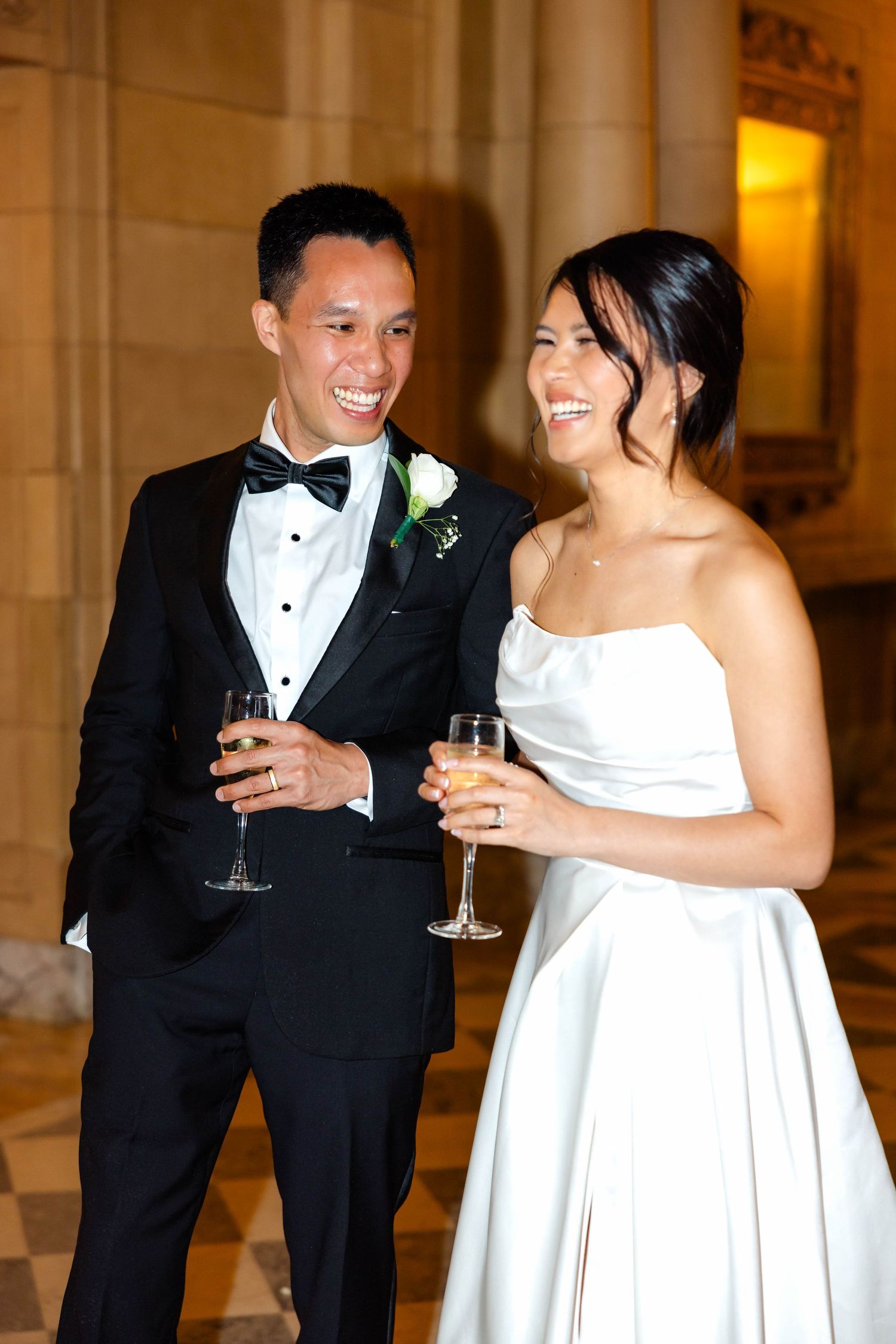 Smiling bride and groom in tuxedo clink champagne glasses in a warmly lit elegant venue.