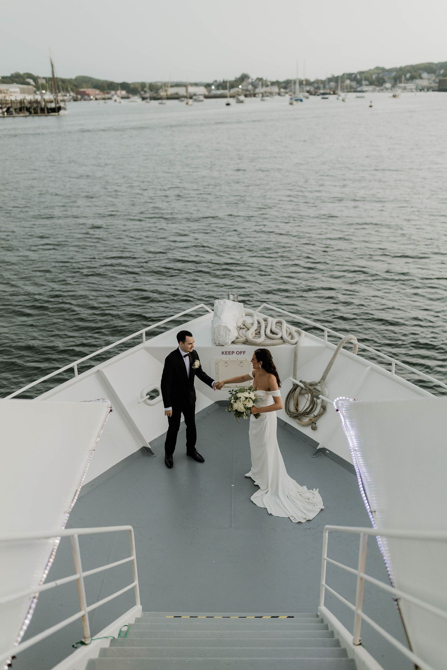 Bride and groom holding hands on bow of boat with scenic harbor backdrop.