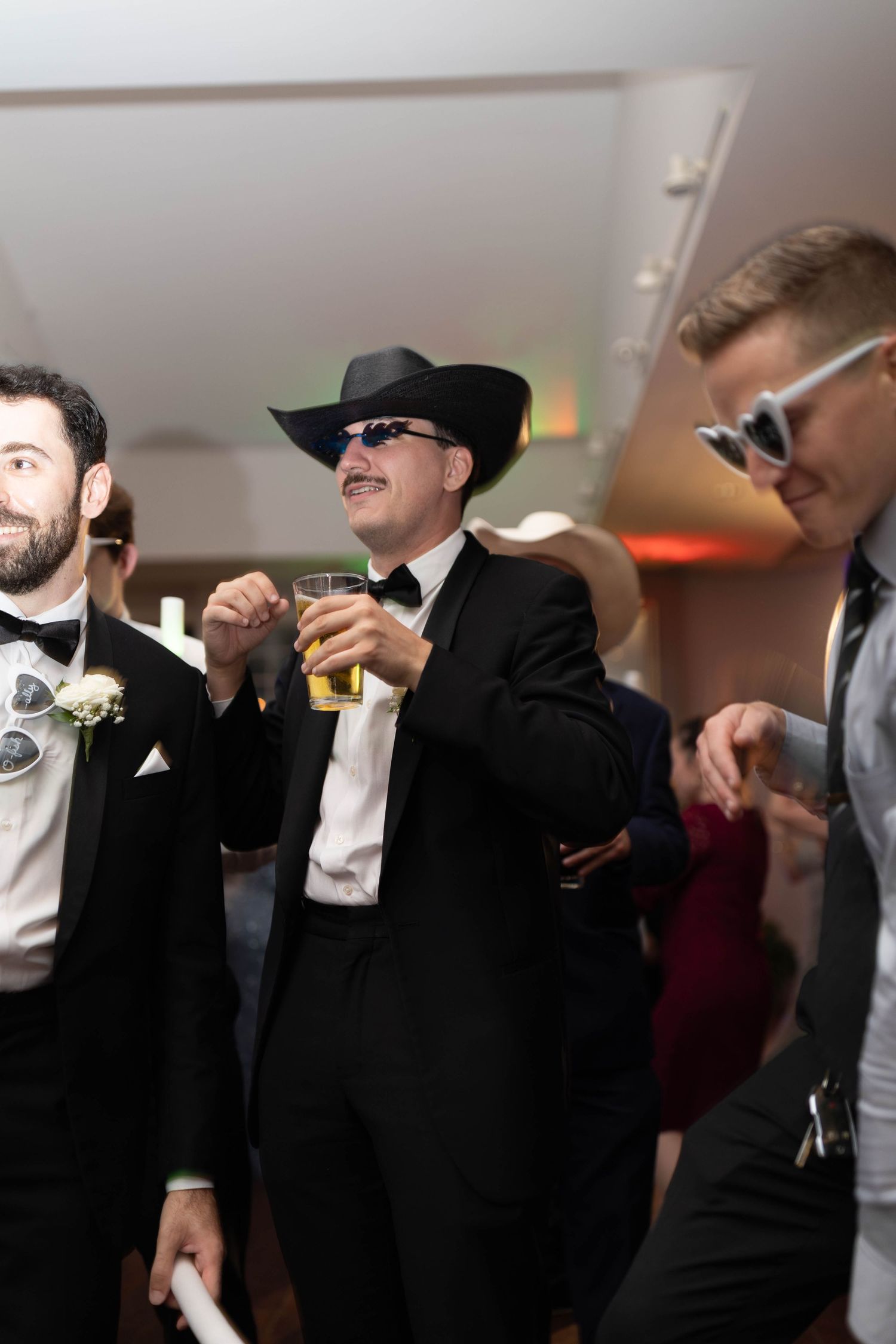 Wedding guests in tuxedos dance wearing cowboy hat and novelty sunglasses at lively reception party.