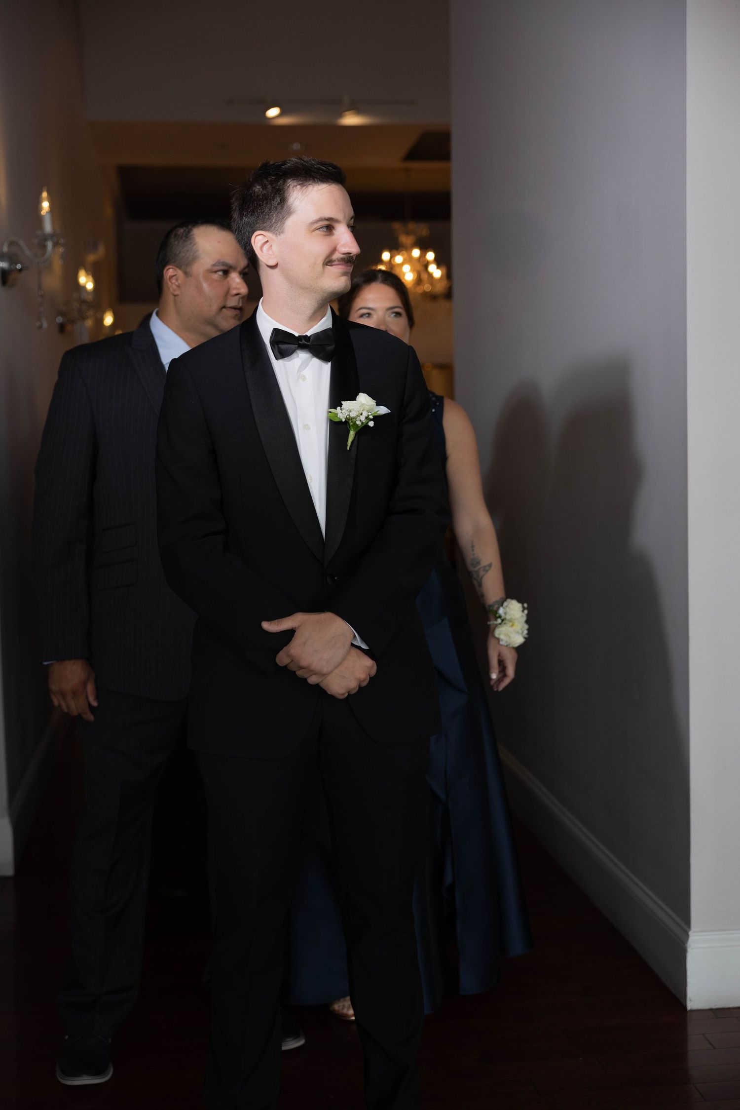Groom in black tuxedo walking with parents before wedding ceremony.