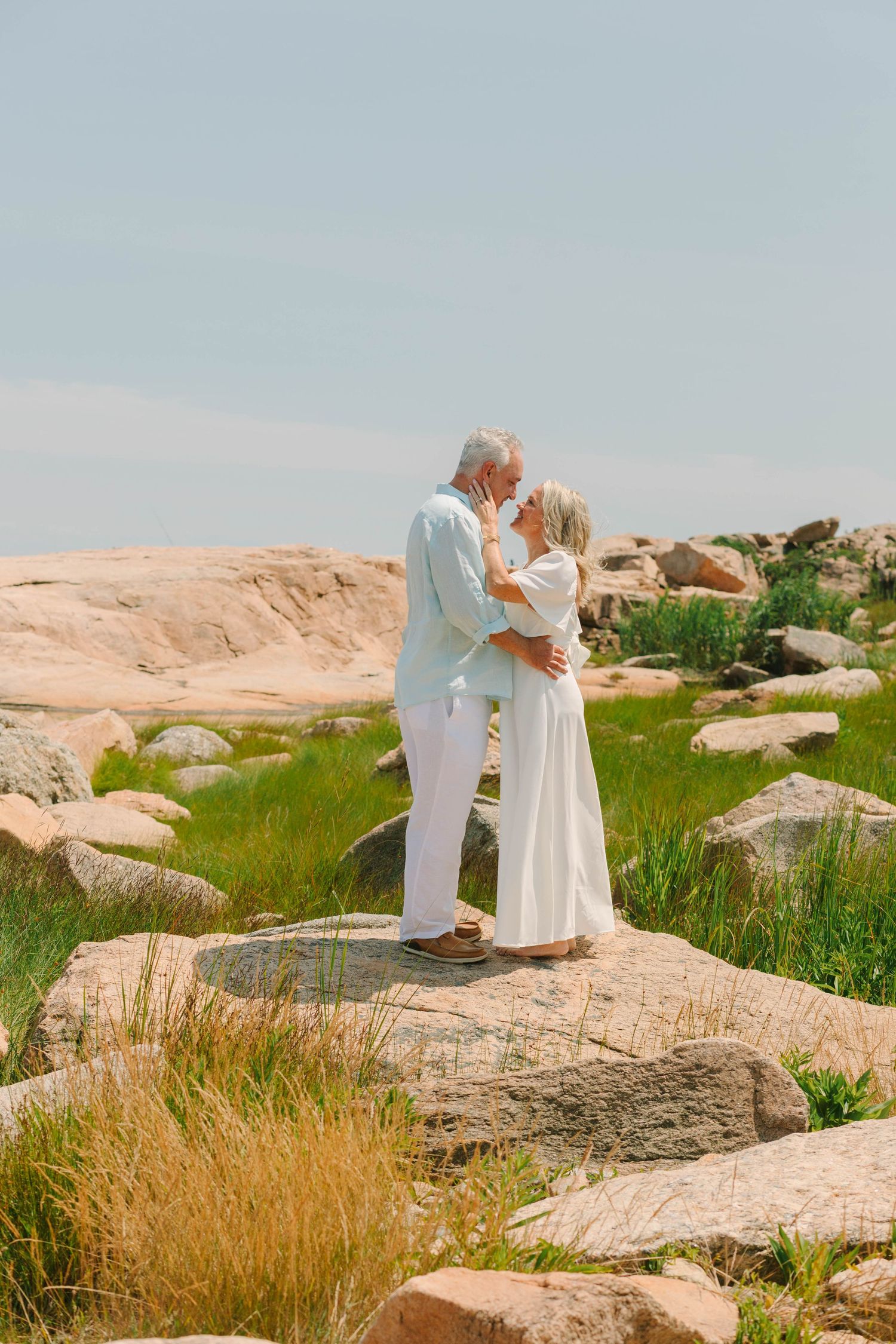 Elderly couple in white attire sharing a romantic moment on rocky coastal landscape with green grass.