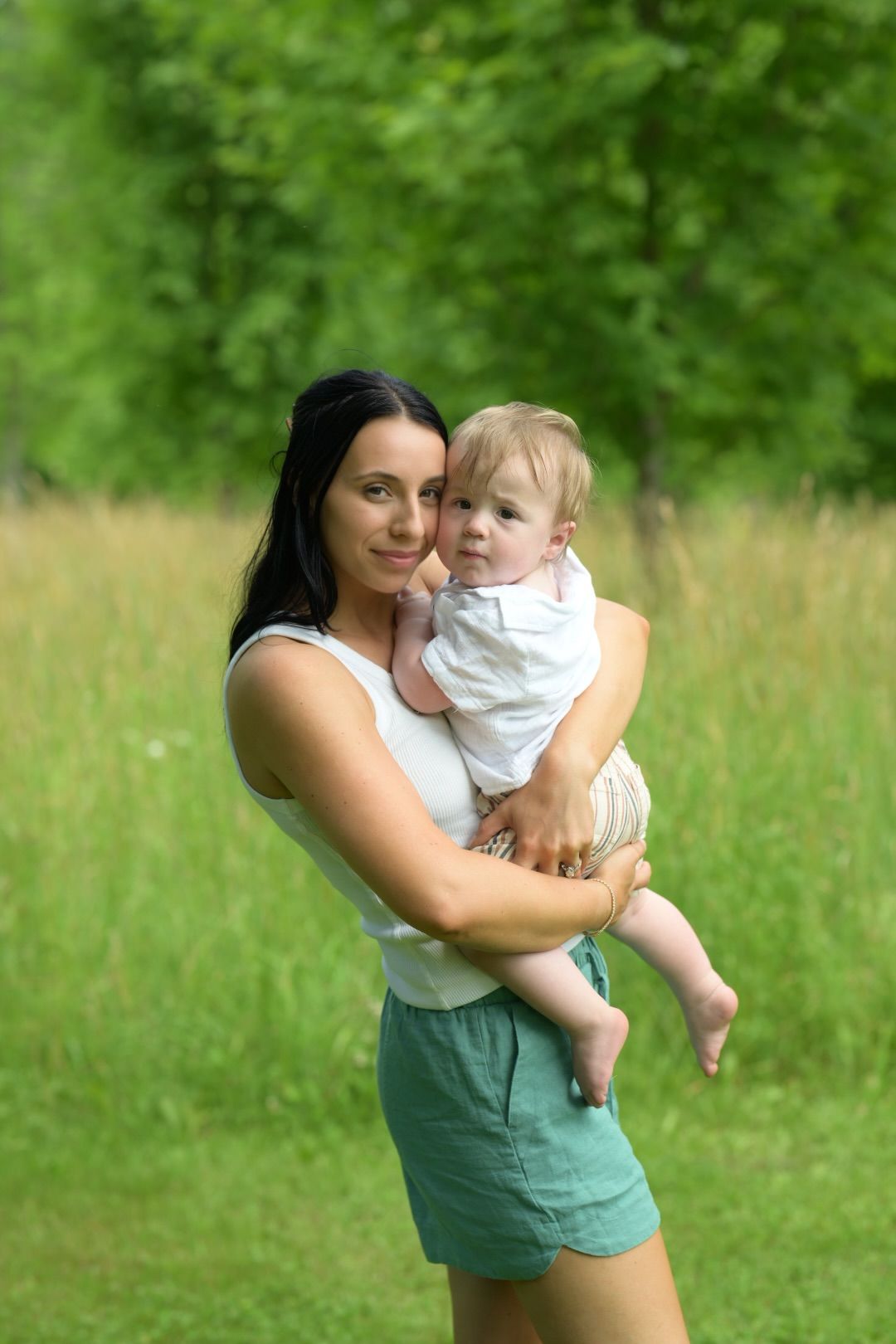 A smiling dark-haired woman holds a cute baby outdoors in a lush green park setting.