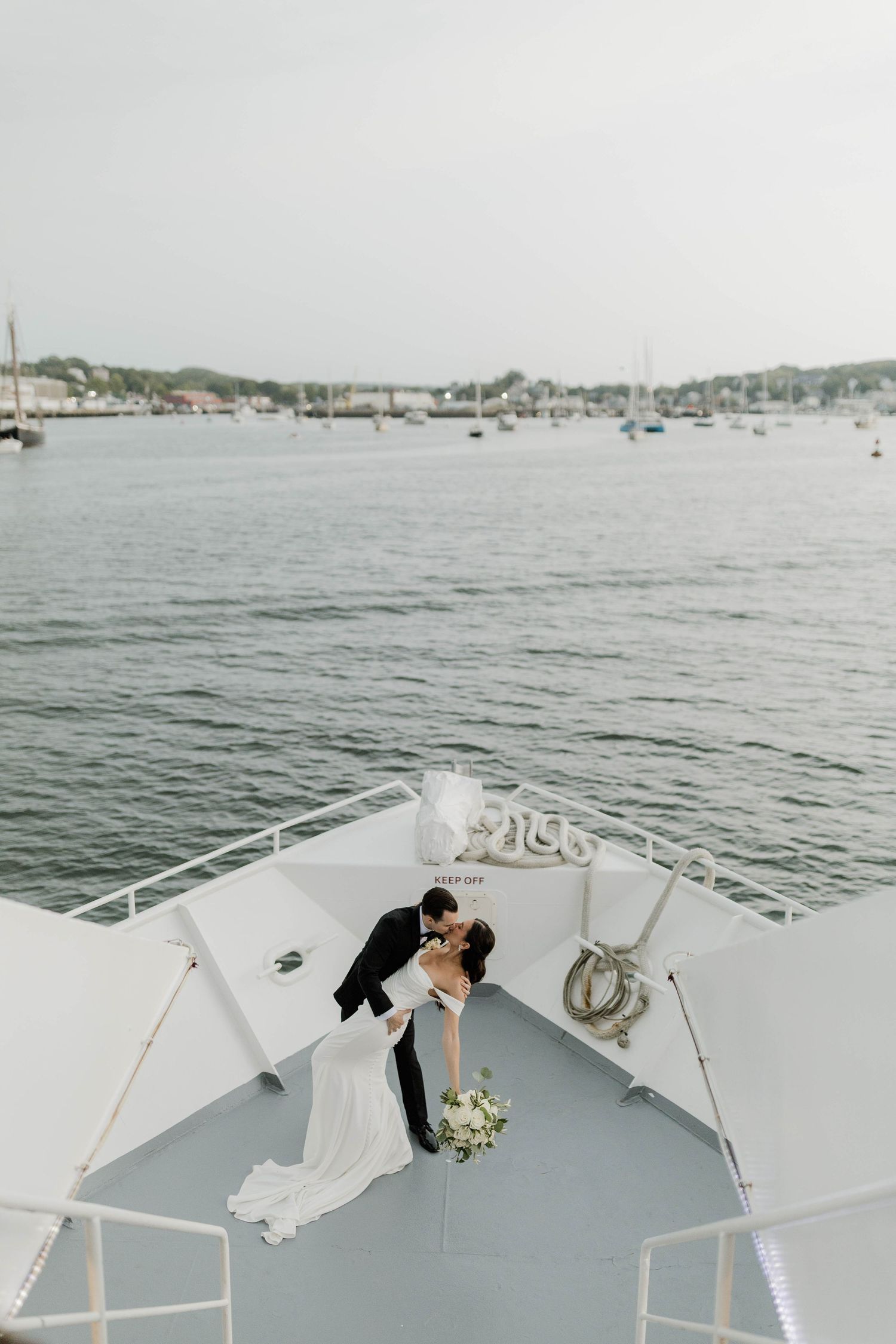 Bride and groom kissing on the bow of a yacht with a scenic harbor and sailboats in the background.