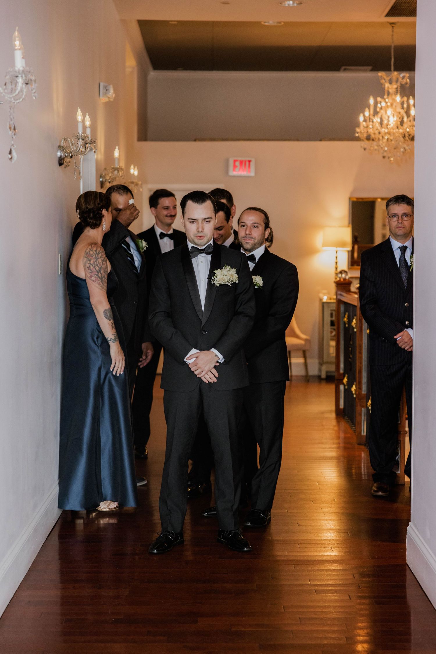 Groom in black tuxedo stands with wedding party in elegant venue hallway before ceremony.