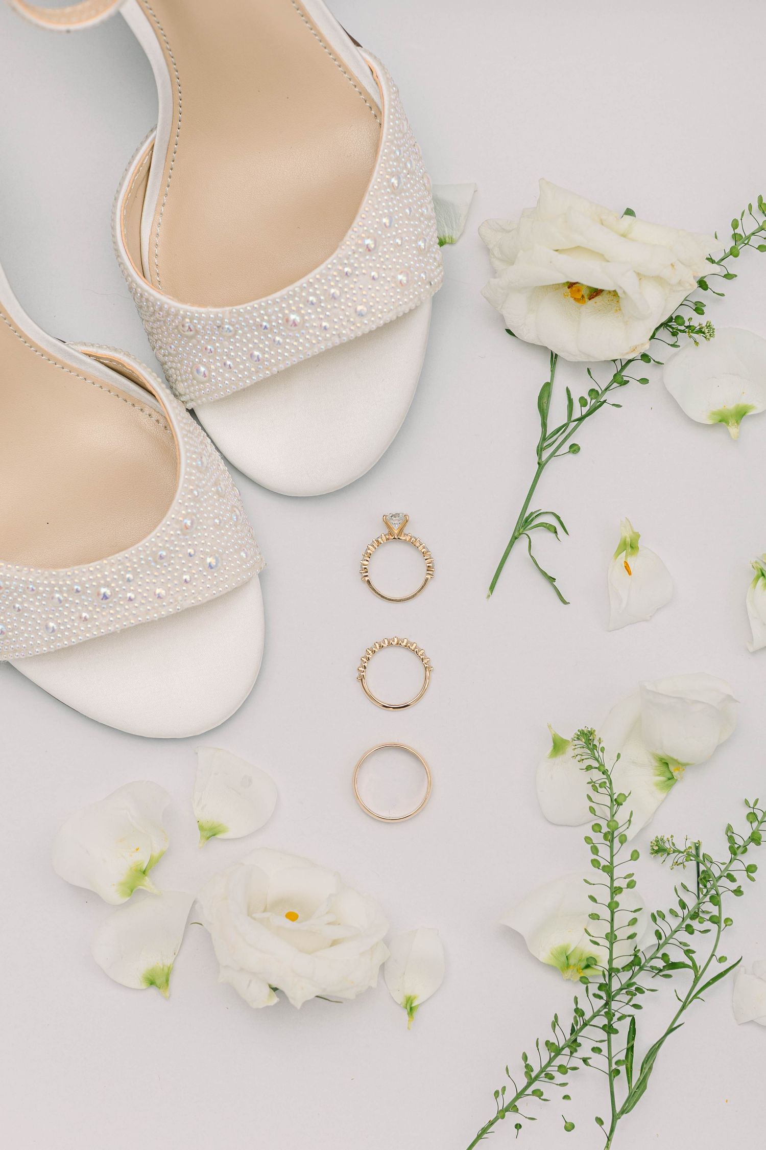 Elegant bridal flat lay featuring pearl-embellished heels, gold wedding rings, and white flowers on a light gray background.