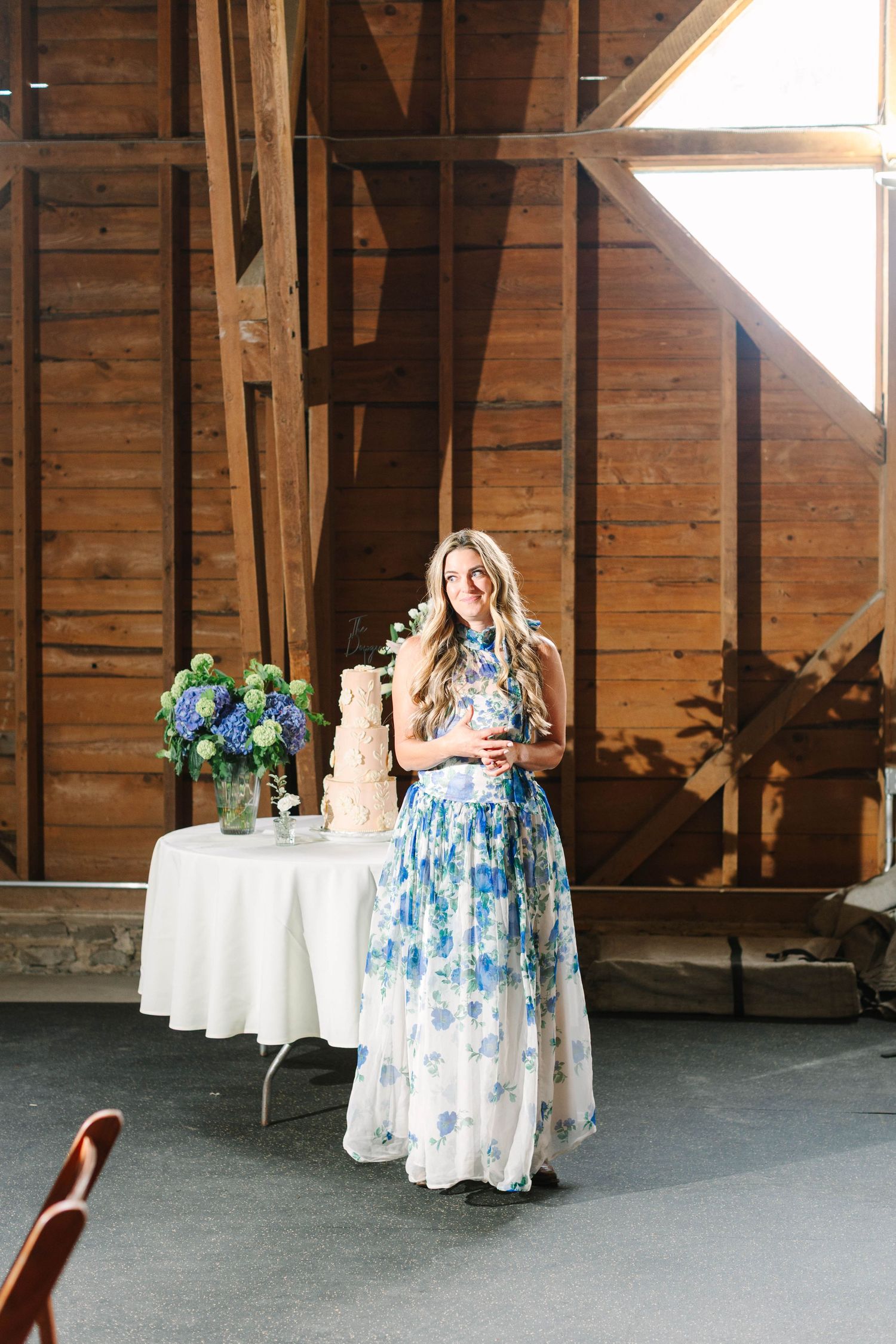 Woman in blue floral gown gives a toast near a wedding cake inside a rustic barn venue.
