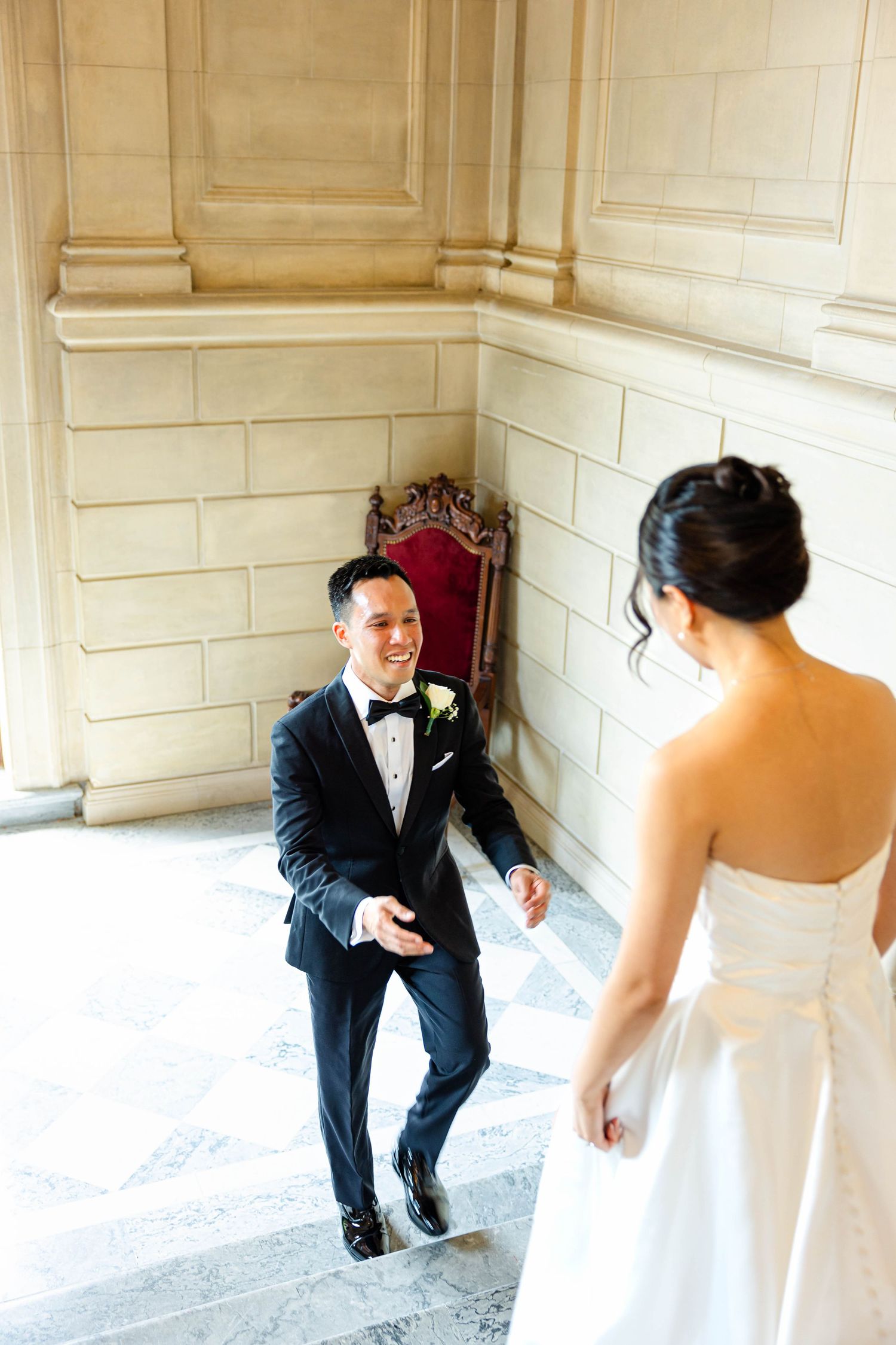 Groom reacts with joyful smile as bride approaches during first look in elegant stone-walled venue.