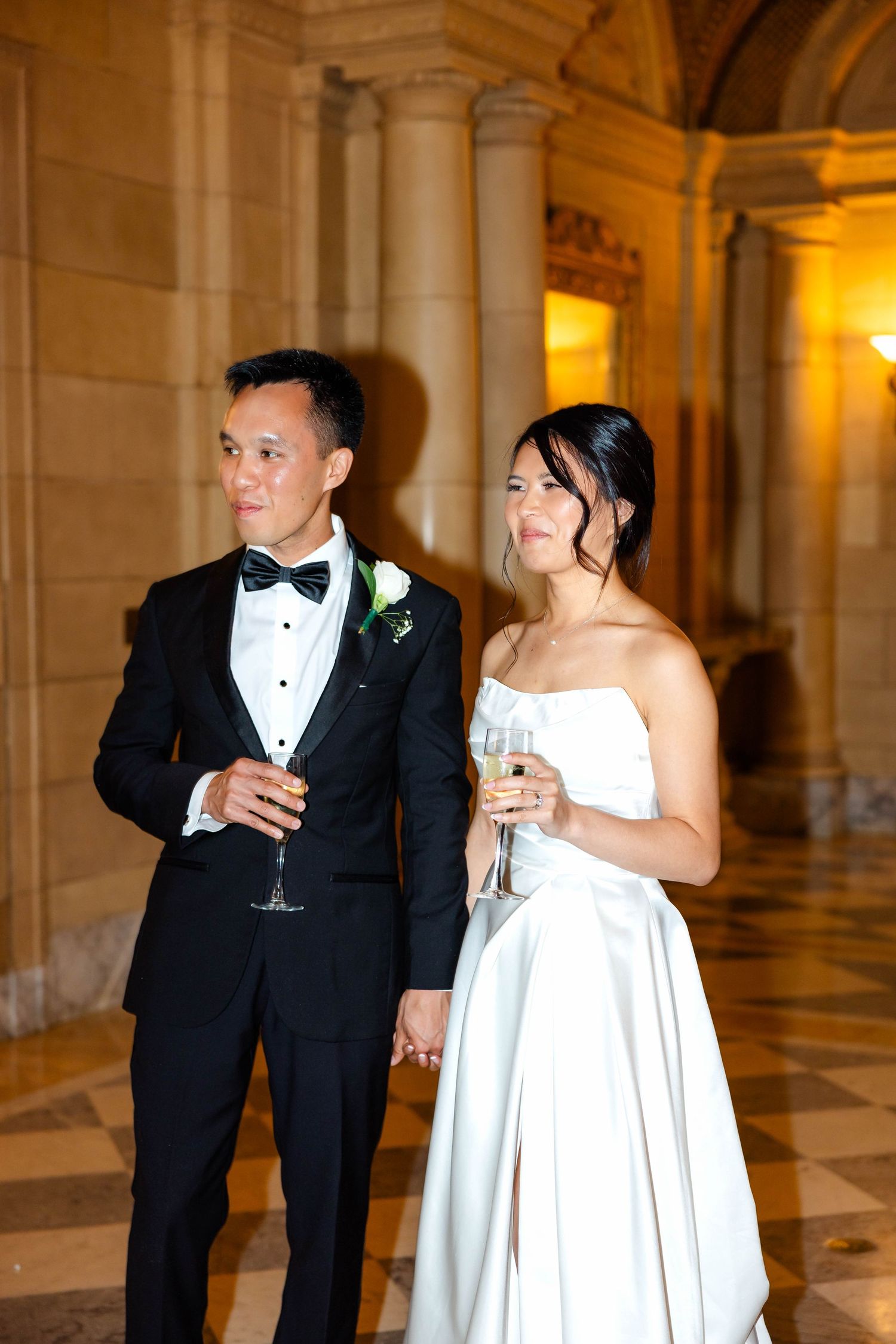 Newlyweds hold champagne glasses hand in hand in ornate golden-lit marble hall at wedding reception.