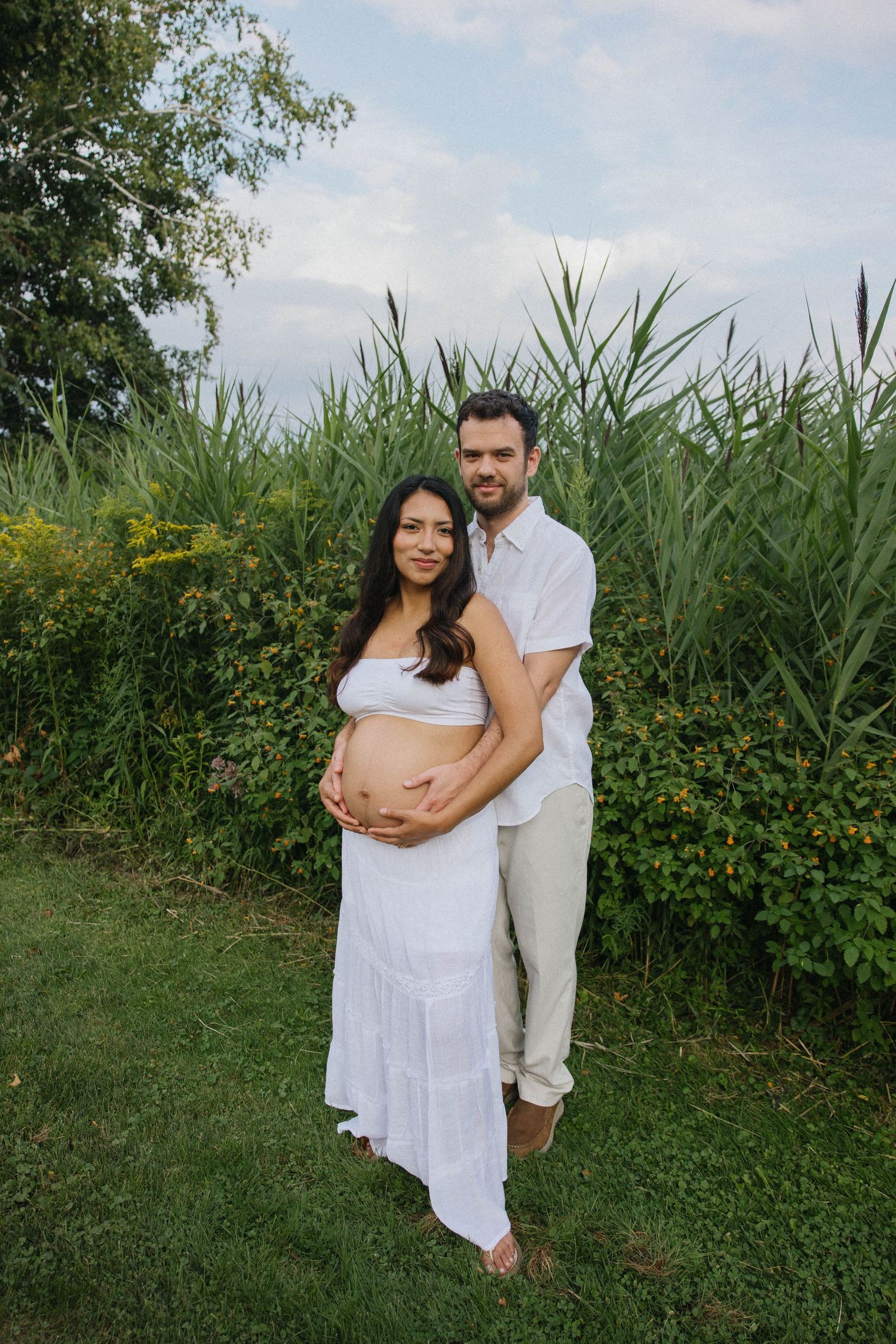 Couple poses outdoors, pregnant woman in white dress with partner embracing her belly near tall green reeds.