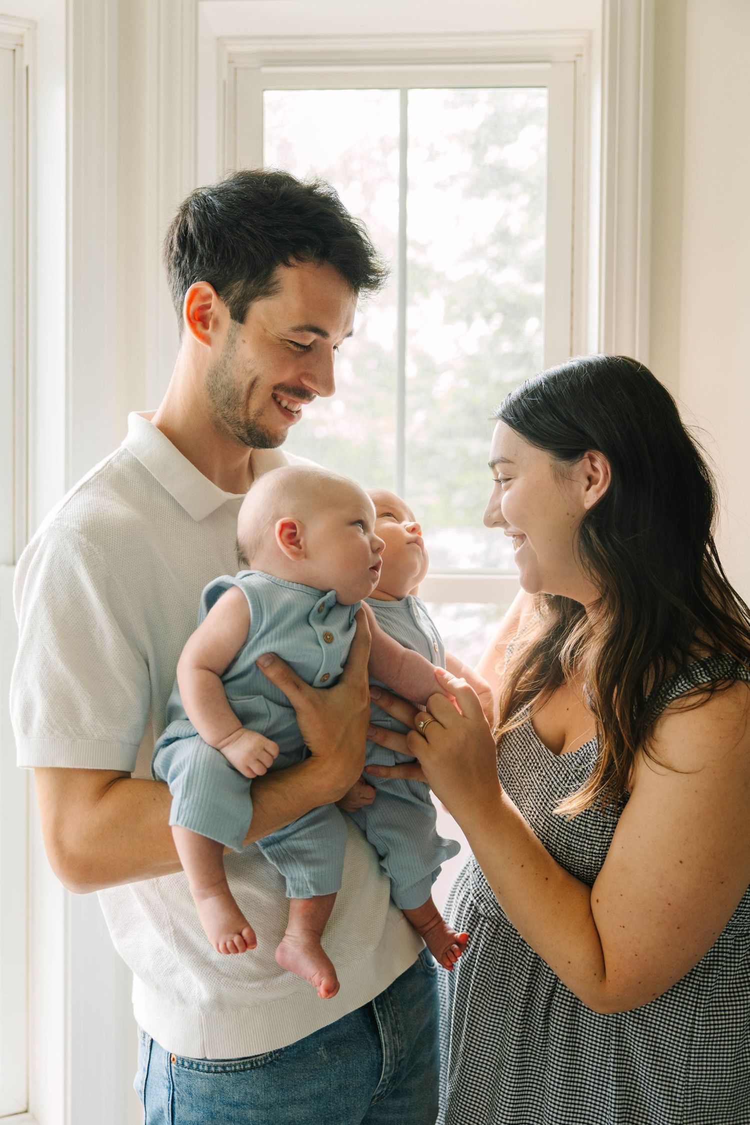 A smiling couple holds their baby in blue overalls near a bright window in a warm family portrait.