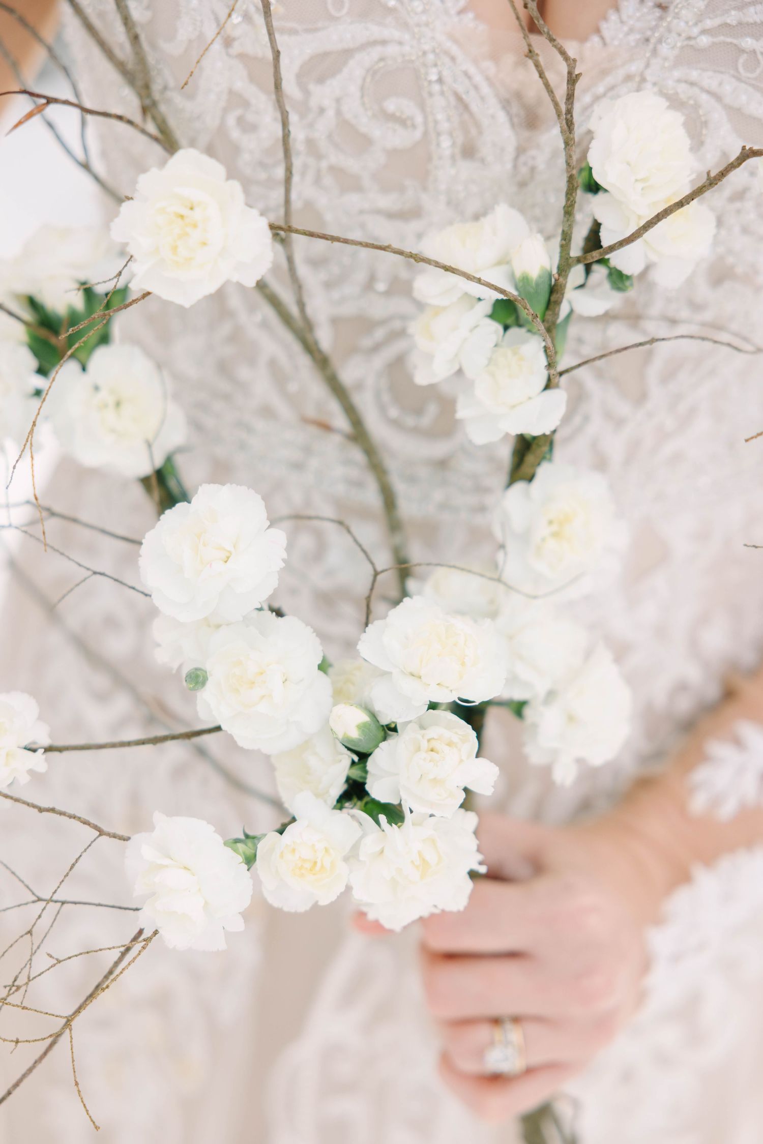Close-up of white carnation wedding bouquet with bare branches held by bride in lace dress.