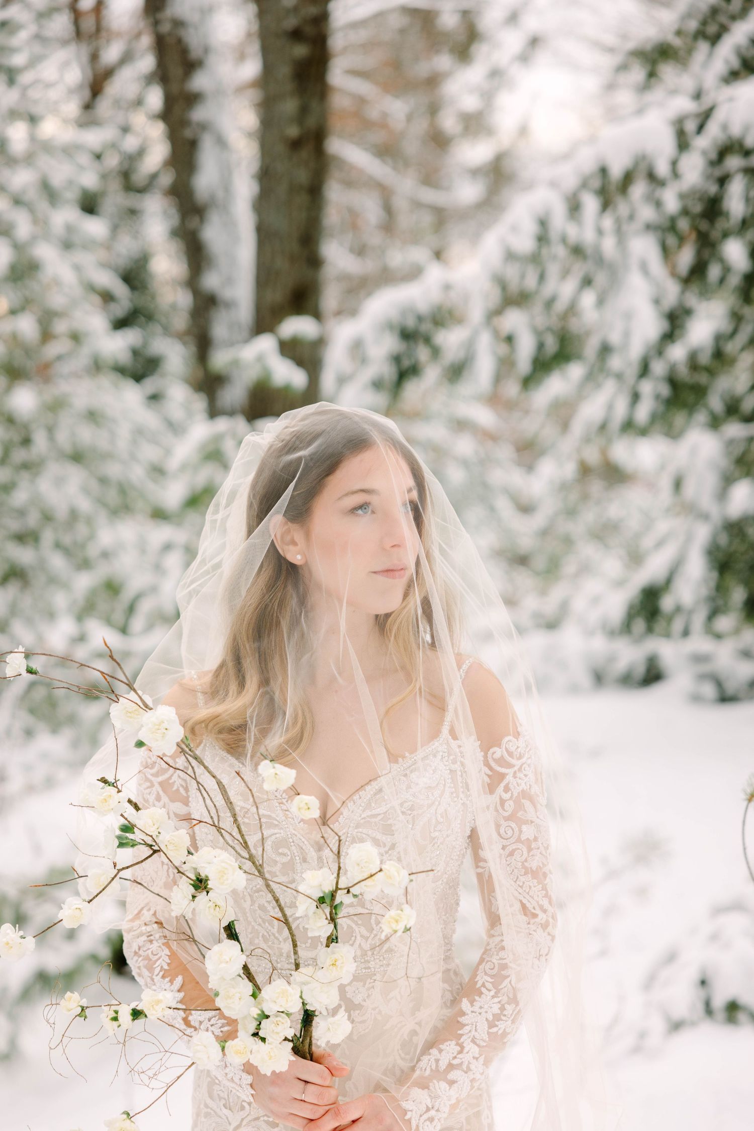 Bride in lace gown with veil gazing aside holding white bouquet in snowy woods.