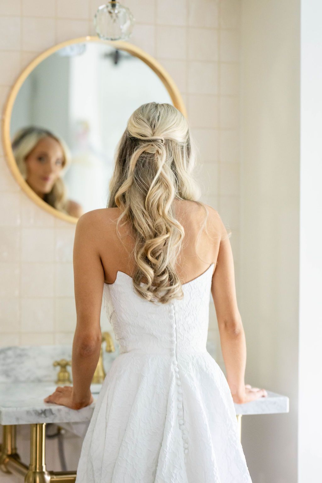 Bride with curly blonde half-up hairstyle in strapless white gown at bathroom mirror.